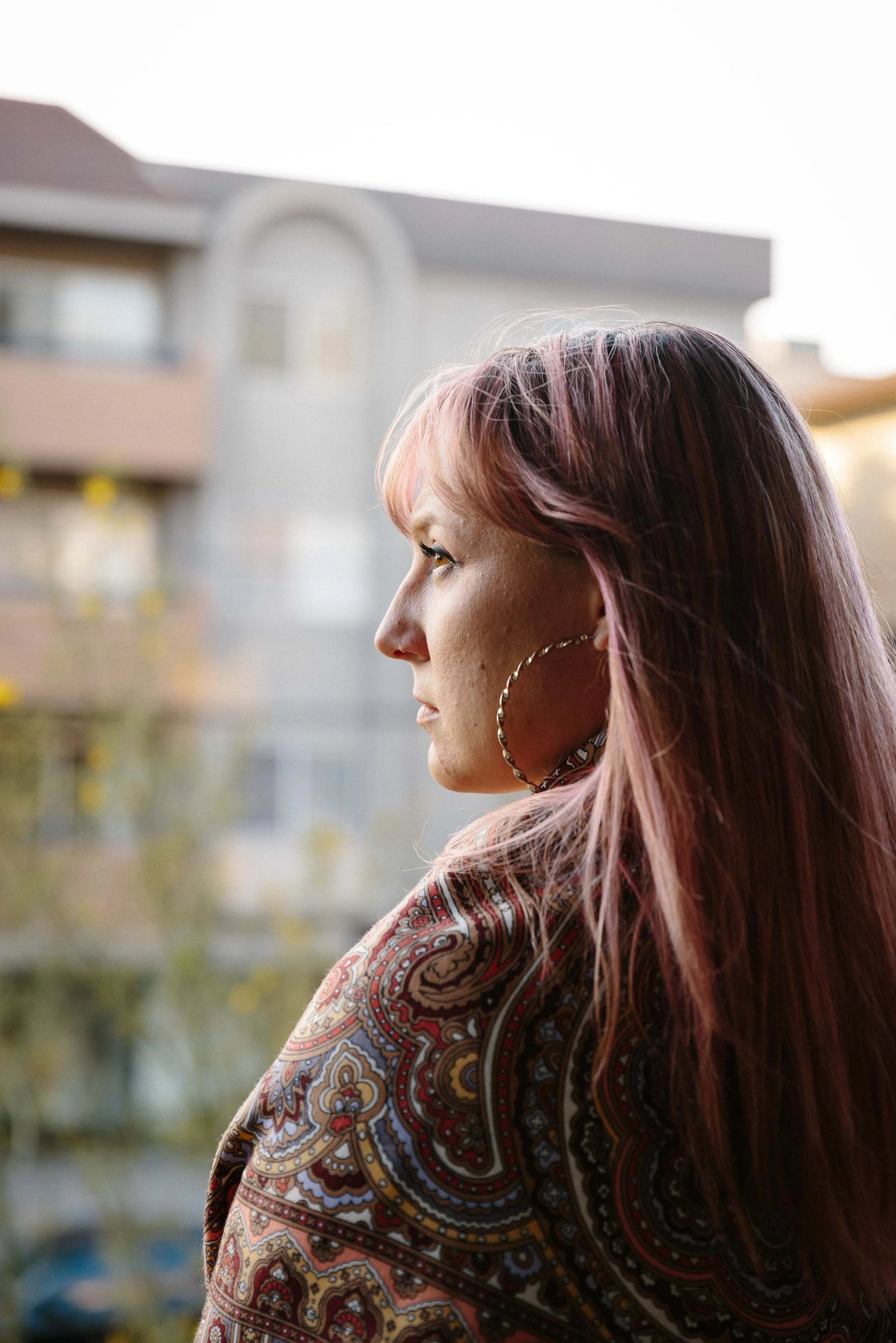 An editorial portrait of a woman photographed in profile with pink hair wearing large hoop earrings and a patterned scarf, looking out of a window with buildings in the background.
