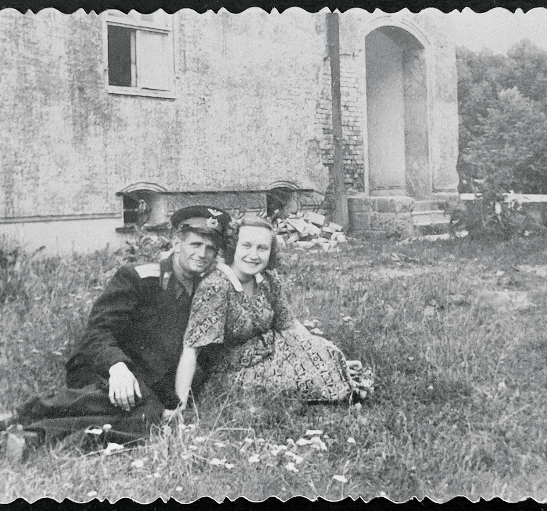 A black and white photo of a man in a uniform and a woman sitting together on the grass outside a house, smiling.