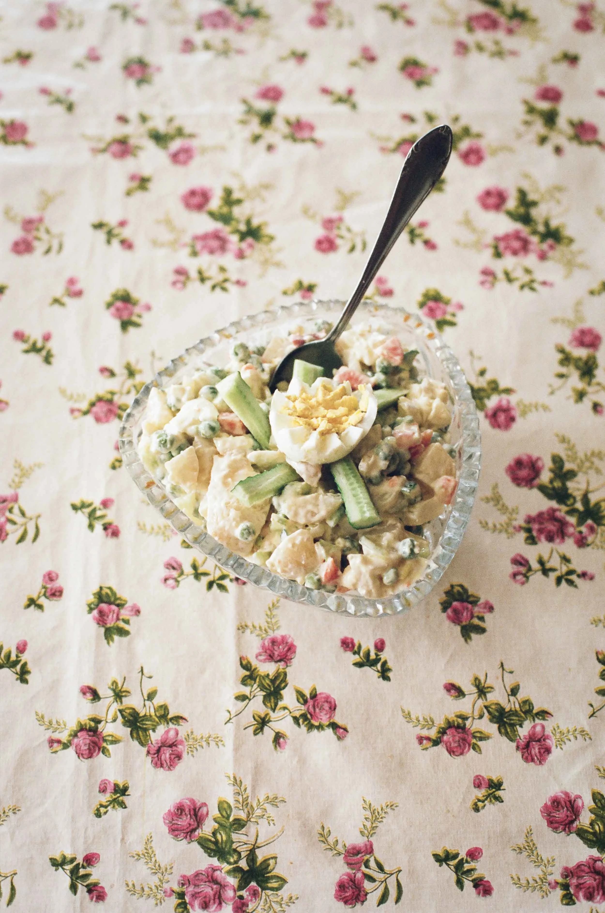 A documentary still life photography of a bowl of egg salad with chopped celery and a sliced hard-boiled egg on top, placed on a floral tablecloth.