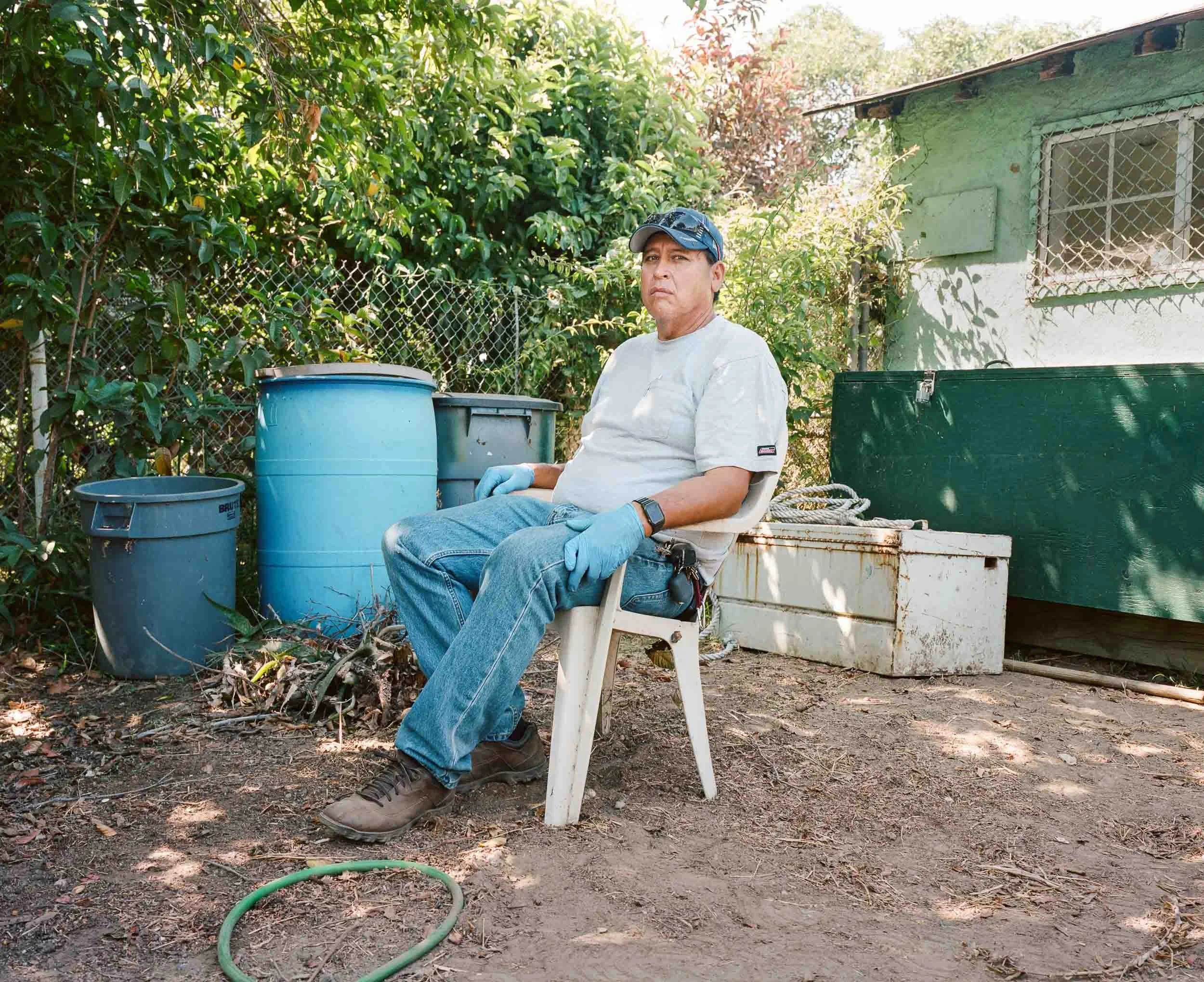 An environmental portrait of a man wearing gloves, a baseball cap, a gray t-shirt, and jeans, sitting on a plastic chair outdoors, with a yard and a green shed behind him. There are various containers and a garden hose on the ground.