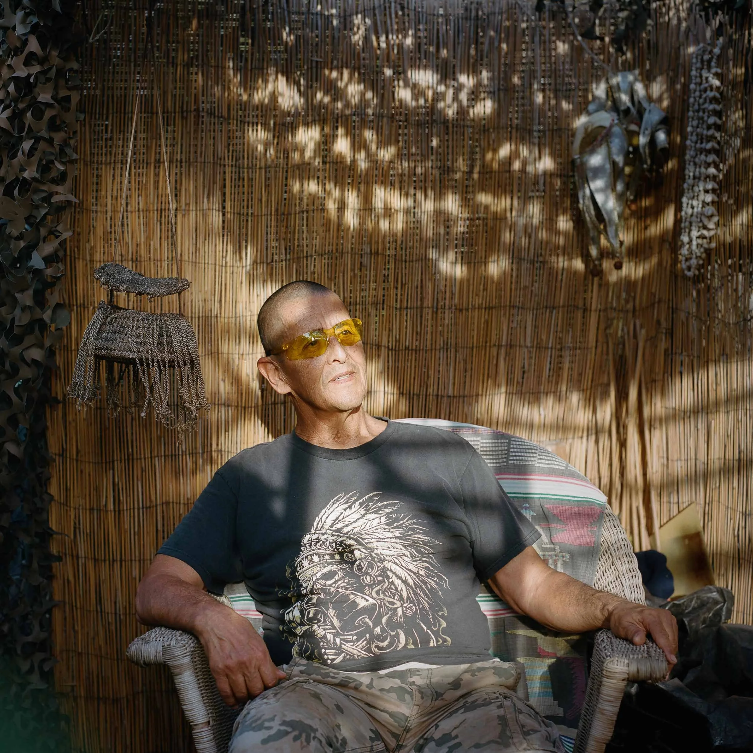 An environmental portrait of an elderly man with a shaved head, wearing yellow-tinted sunglasses and a black T-shirt with a lion graphic, sits in a wicker chair against a bamboo backdrop.