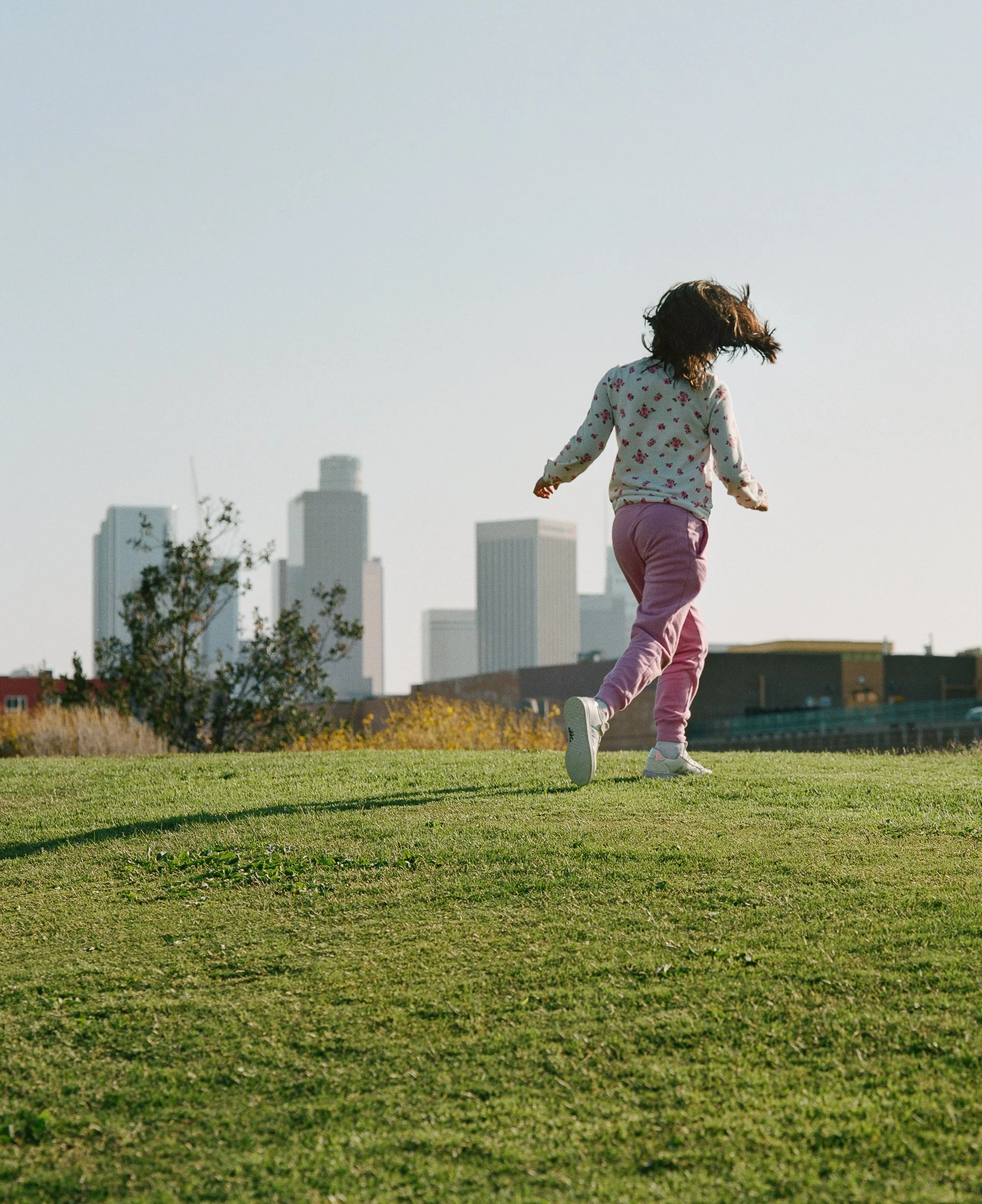 An editorial photograph of a girl running on a grassy hill with a city skyline in the background.