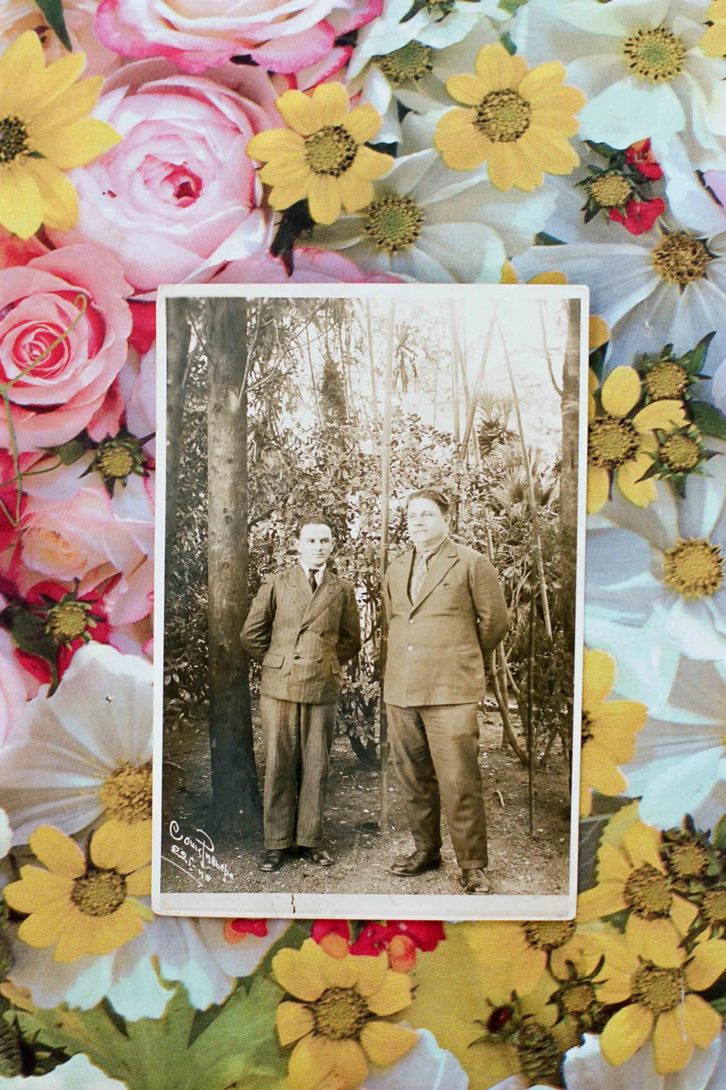 A detail photograph of a black and white photograph of two men in suits standing outdoors among trees and plants. The photo is placed on a colorful floral background with pink, yellow, and white flowers.
