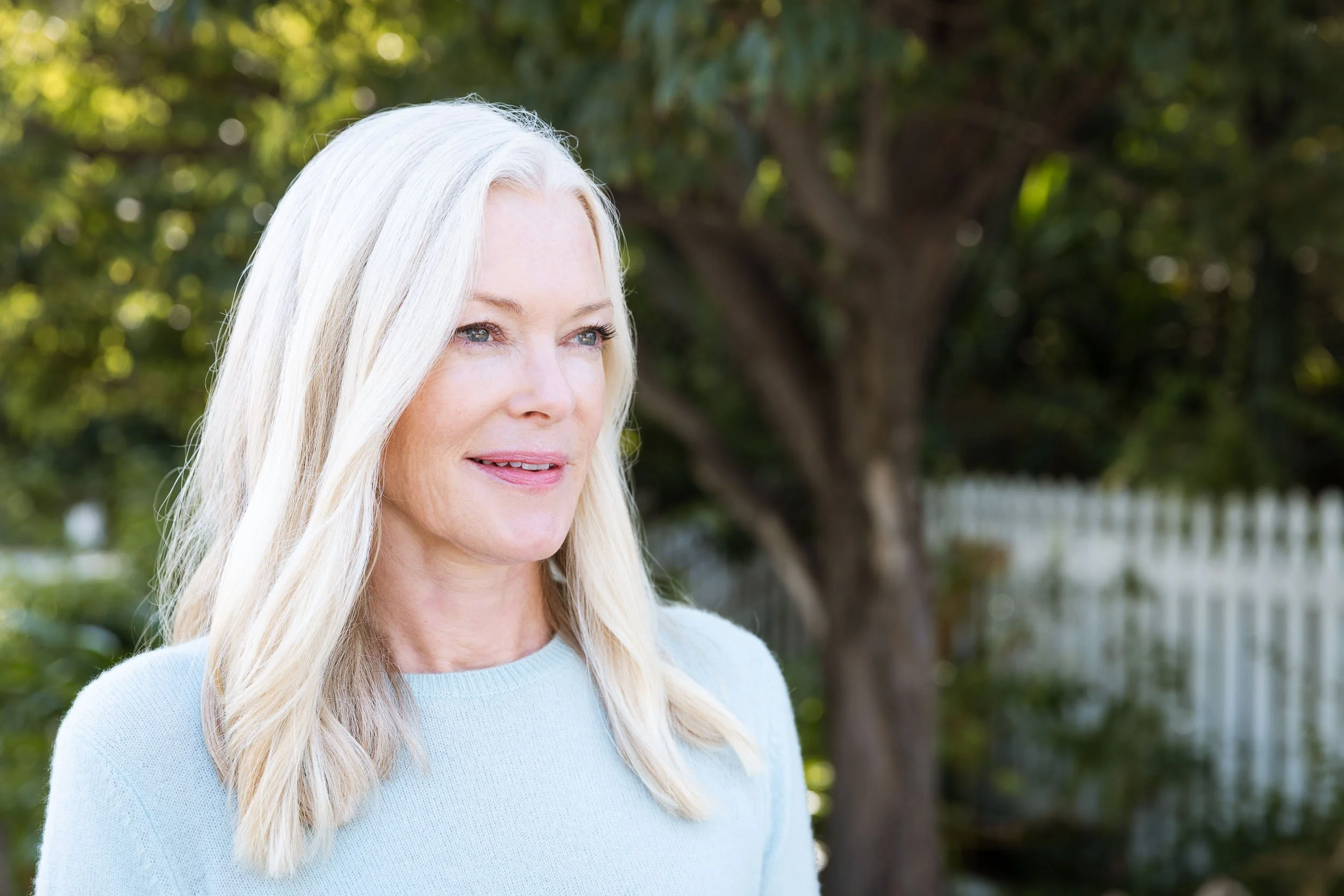 A closeup portrait of a woman with long blonde hair wearing a light blue sweater outdoors with a tree and white fence in the background.