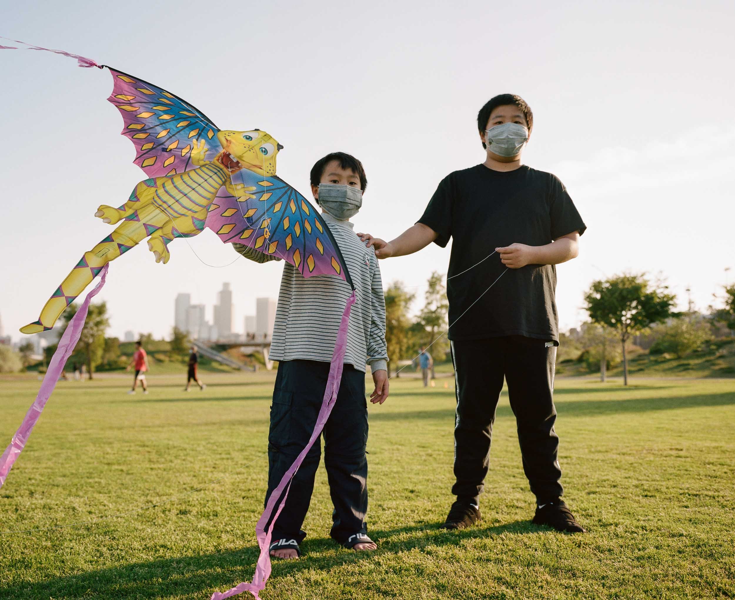 An environmental potrait of two boys wearing face masks in a park, one holding a colorful butterfly-shaped kite, with trees and a city skyline in the background during sunset.