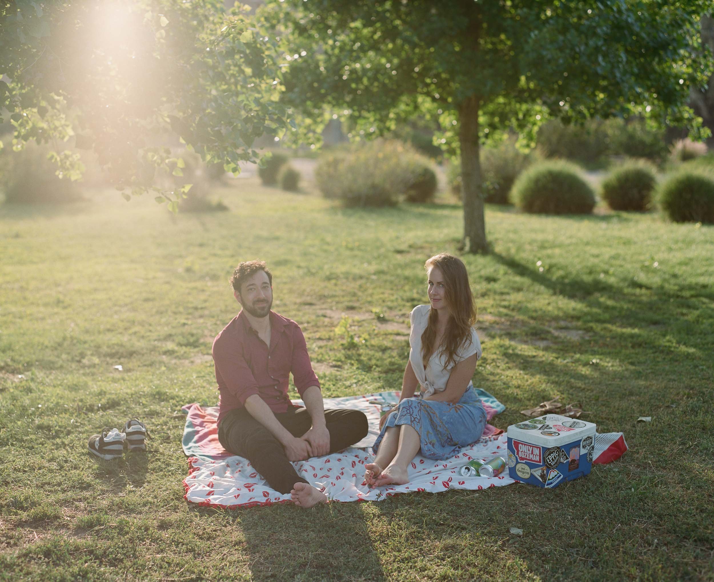 An editorial portrait of a man and a woman sitting on a blanket during a picnic in a park, with trees and bushes in the background, and sunlight shining through the leaves.