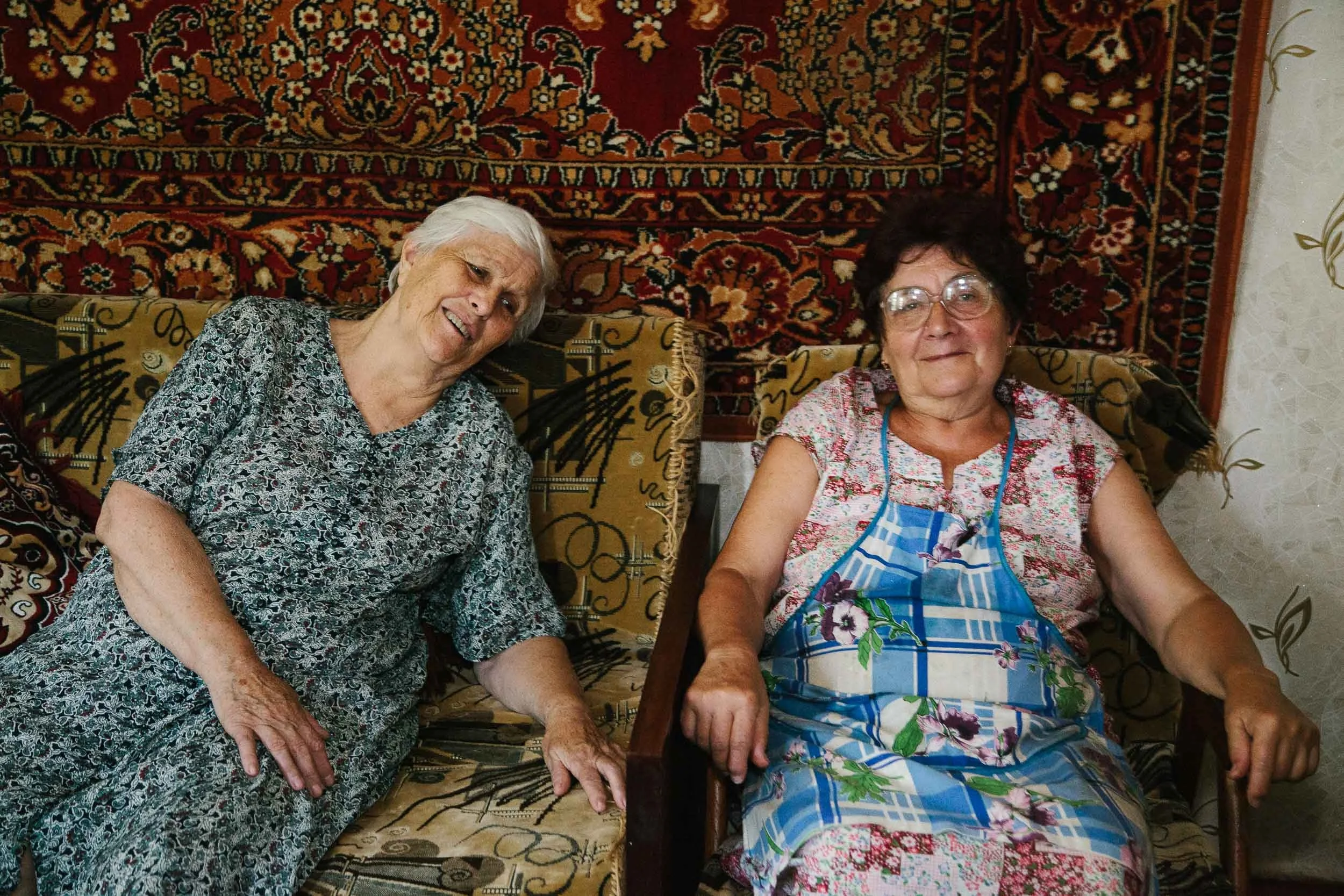 Two elderly women sitting on a sofa in a cozy living room, smiling at the camera. One woman has white hair and wears a patterned dress, while the other has dark hair, glasses, and wears a floral dress with an apron. A decorative patterned rug hangs on the wall behind them.