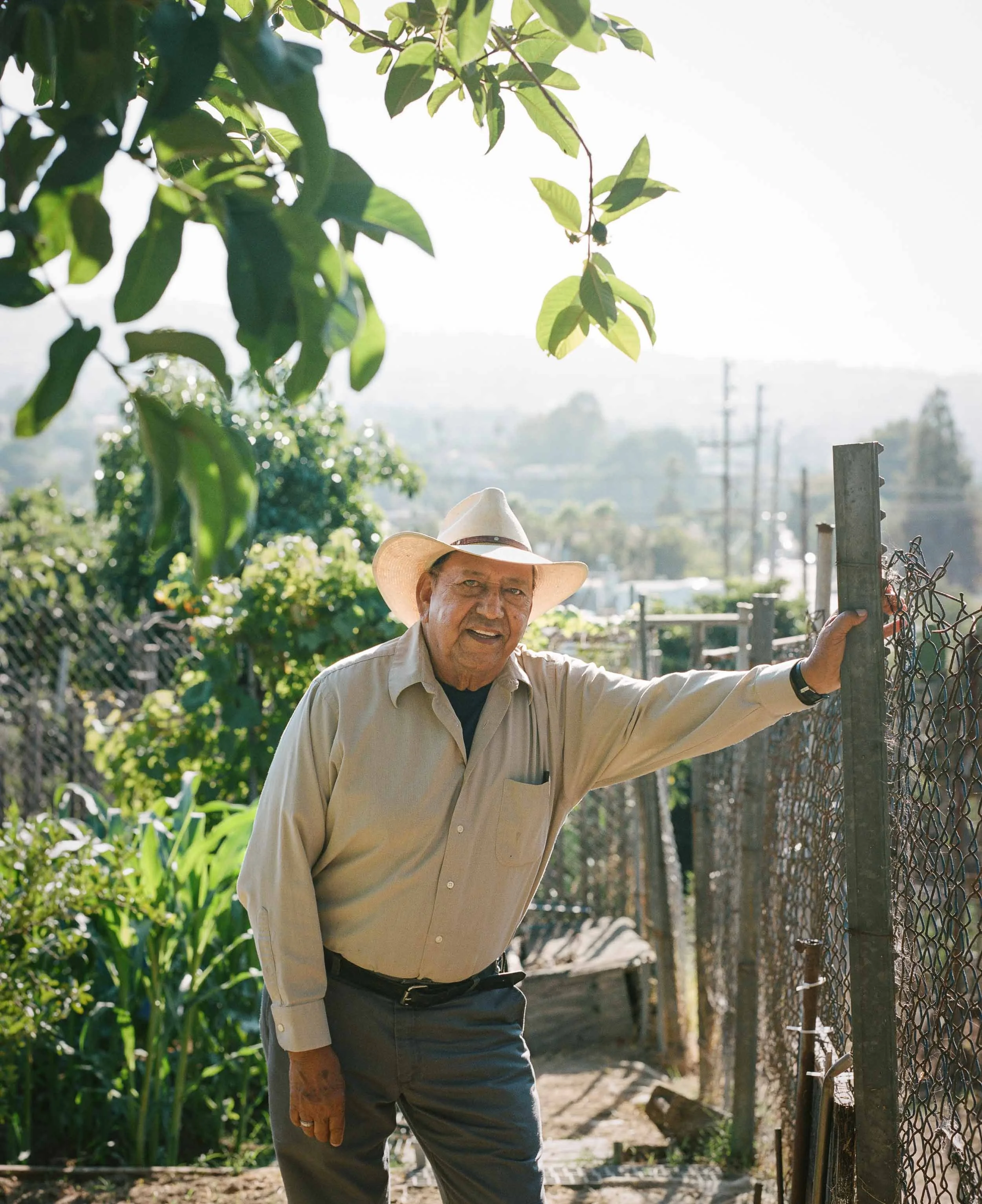 An environmental portrait of an elderly man wearing a cowboy hat and a beige button-up shirt stands outdoors, holding onto a chain-link fence with a rural landscape in the background.