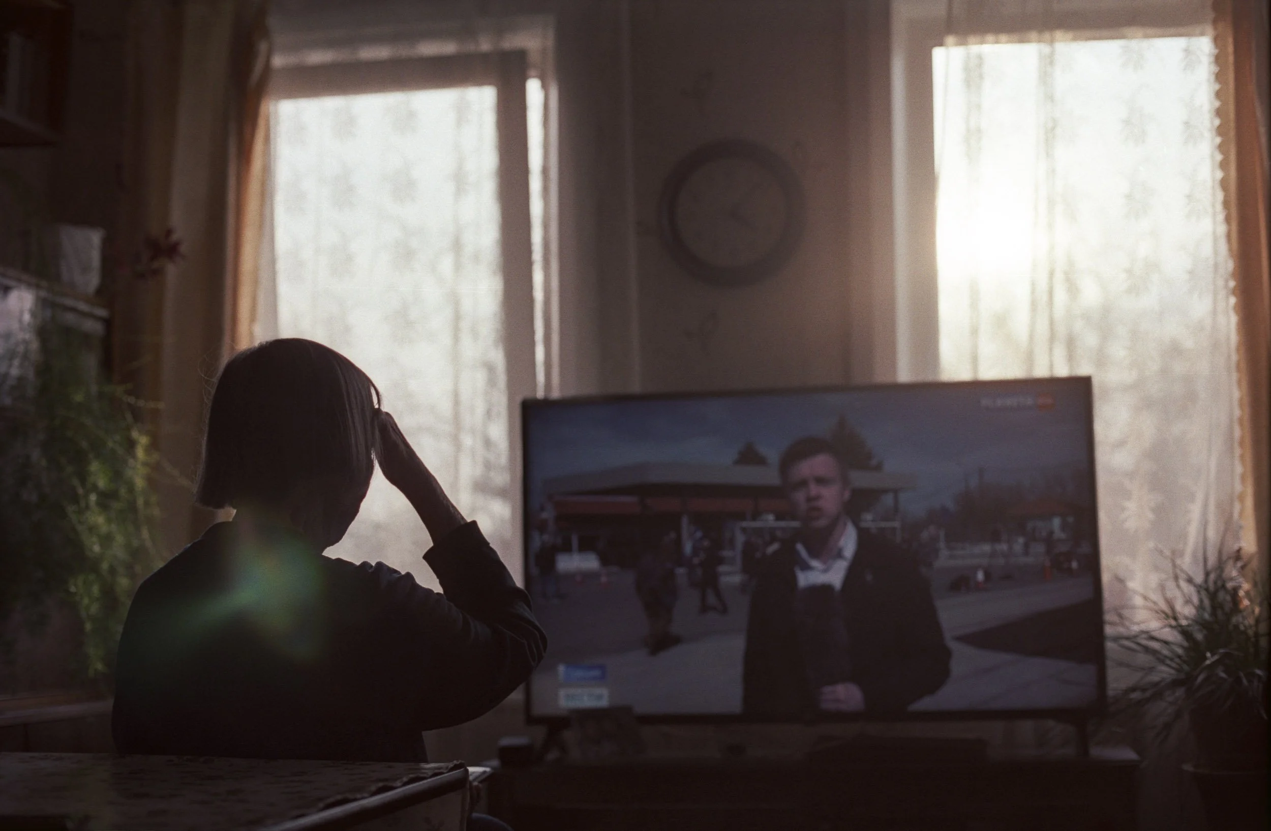 A documentary photograph of a person sitting at a table watching television in a room with sunlight coming through the windows.