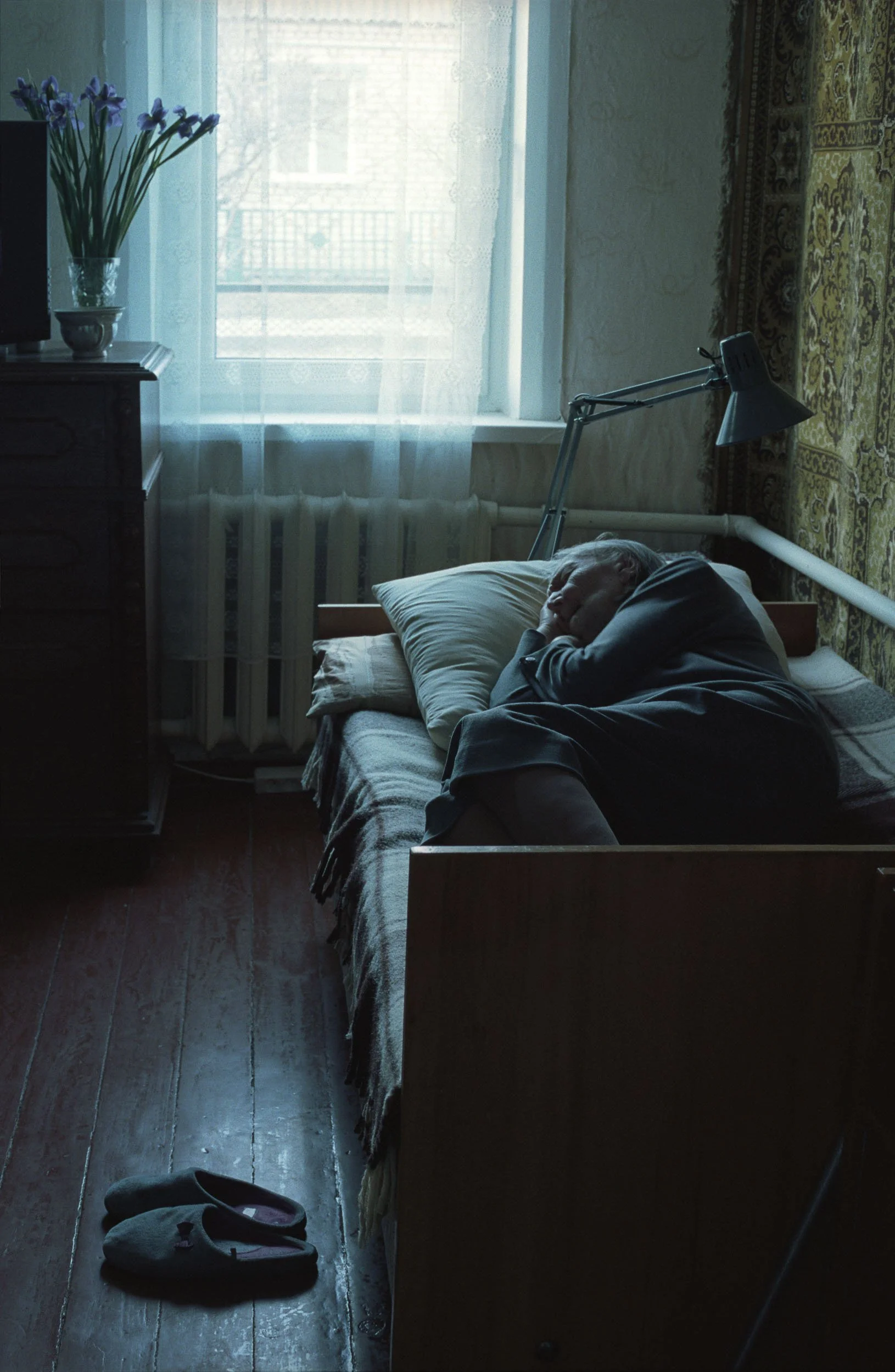 A documentary photograph of an elderly person sleeping in a bed in a dimly lit room with a window, a vase of flowers, and a lamp.