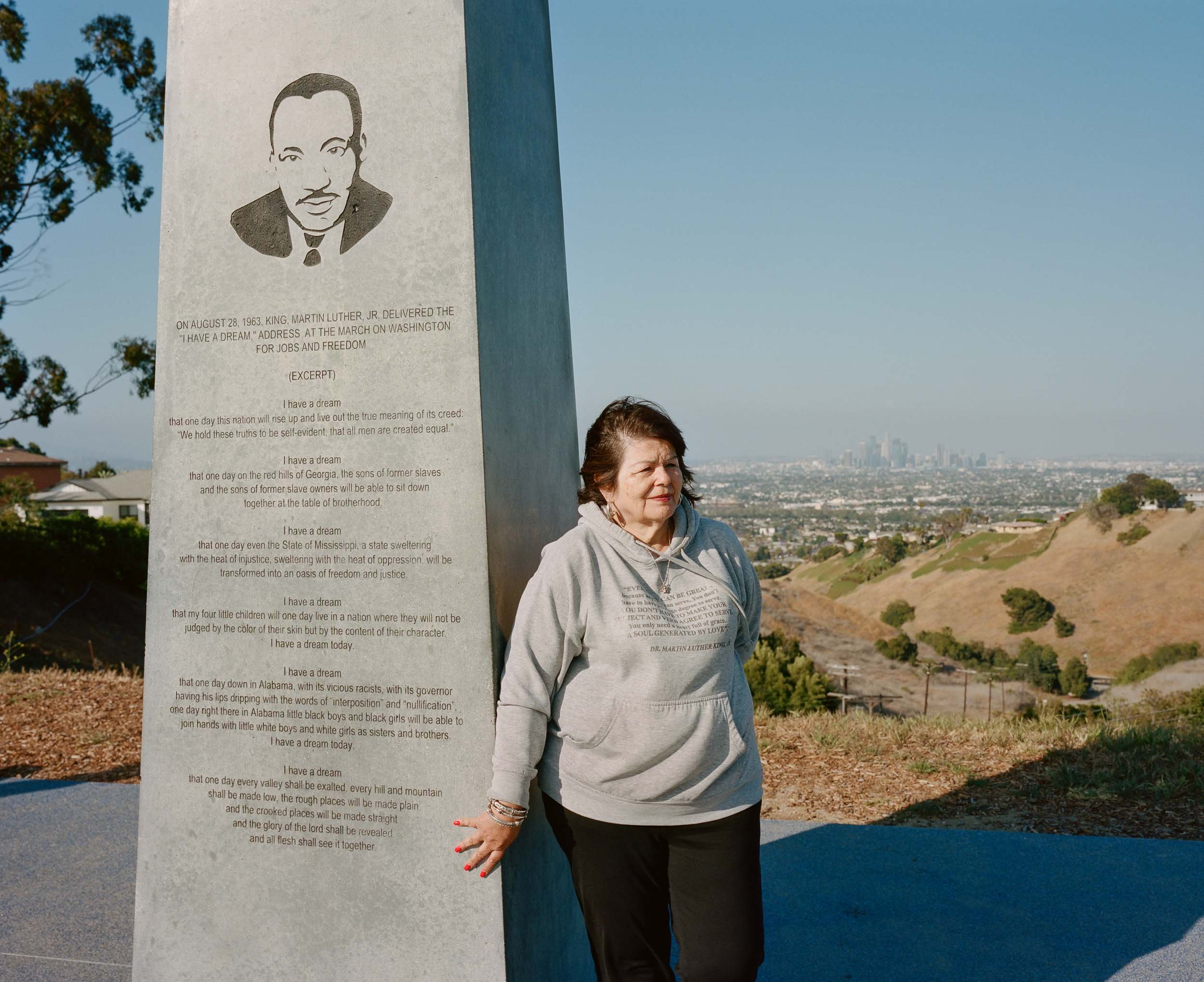 An environmental portrait of a woman standing outdoors next to a tall, gray monument with an illustrated portrait and a quote by Dr. Martin Luther King Jr. The monument features his image and an excerpt from his speech.