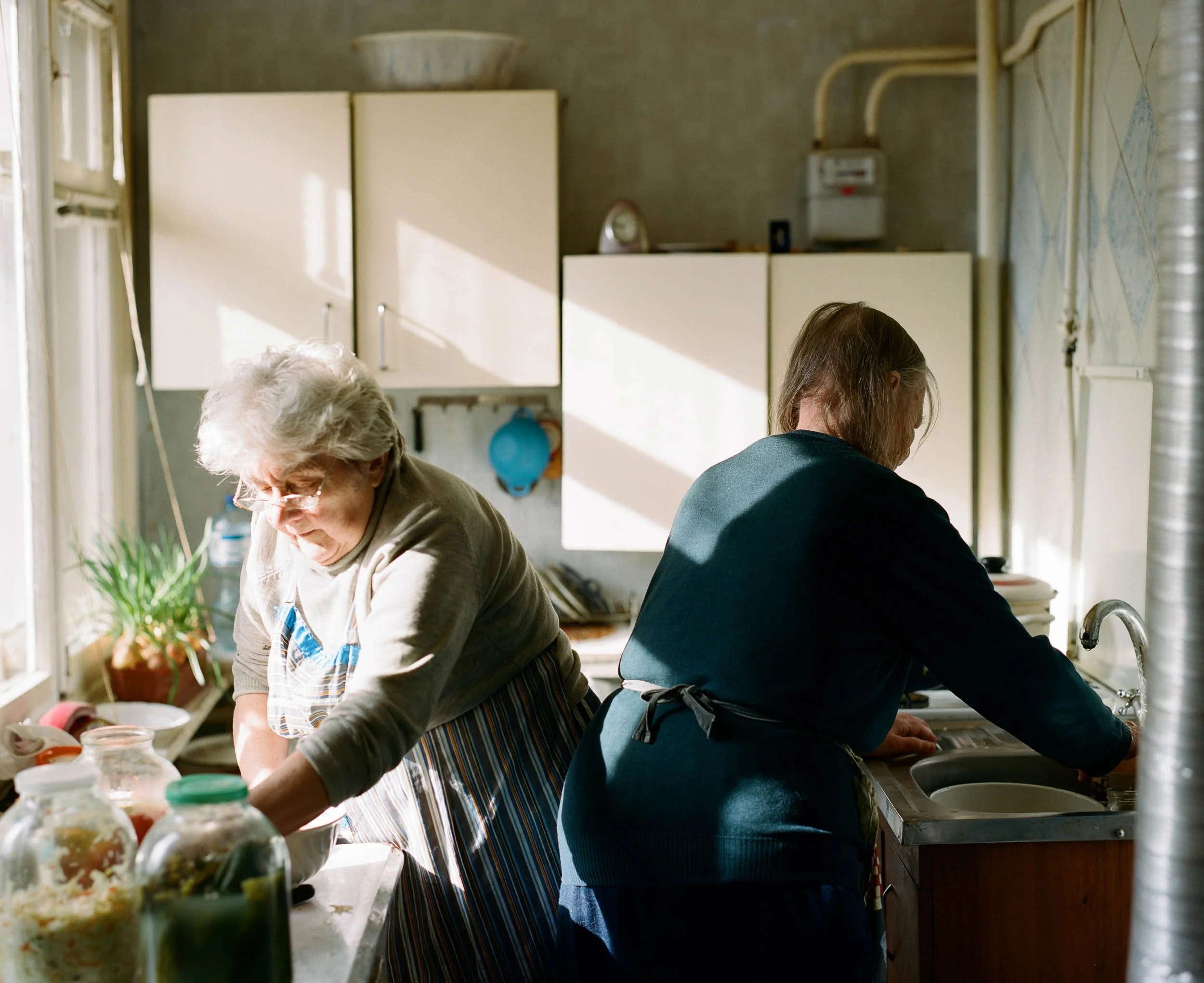 A documentary photograph of two women are washing dishes in a kitchen with sunlight streaming through the window.