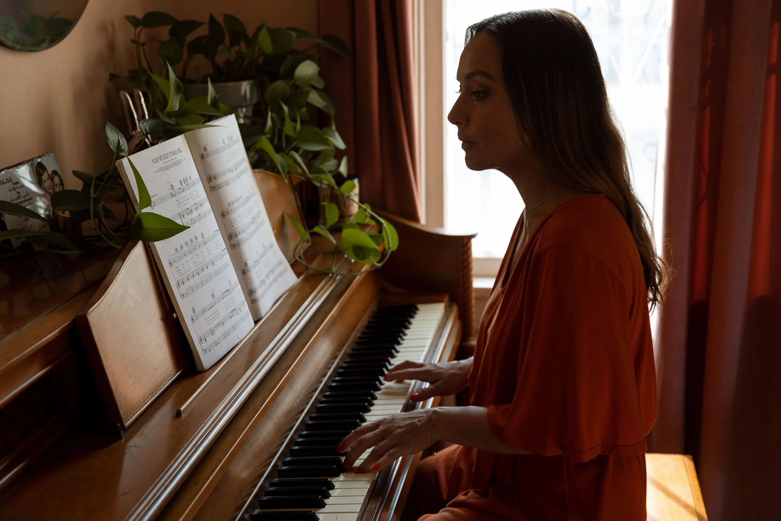 A lifestyle photograph of a woman playing the piano in a room with sunlight coming through the window, placed beside plants and sheets of music.