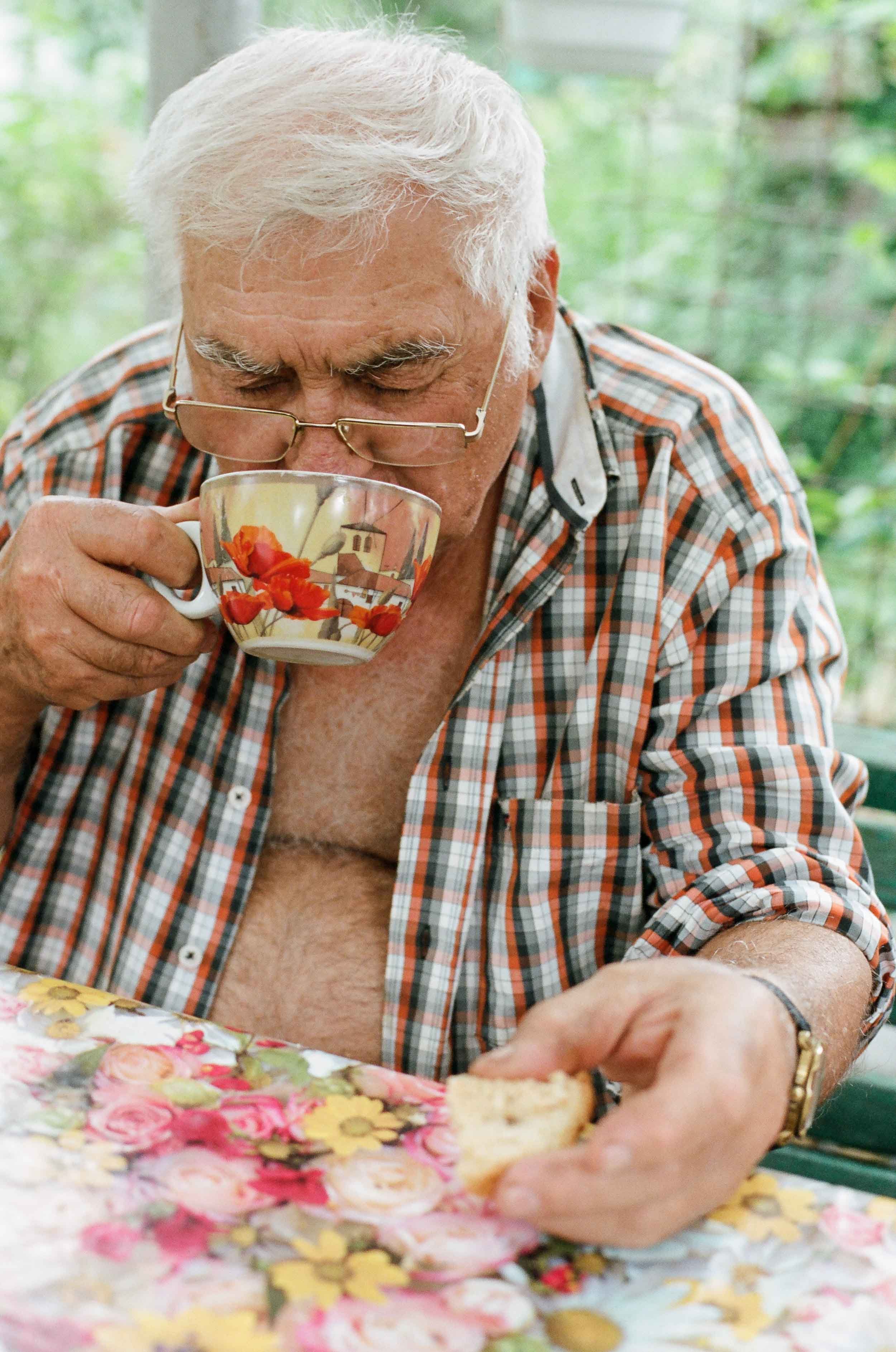 A elderly man with gray hair, wearing glasses and a plaid shirt, is drinking from a floral teacup while holding a biscuit in his other hand. He is seated at a table with a colorful floral tablecloth, in a setting with green foliage in the background.
