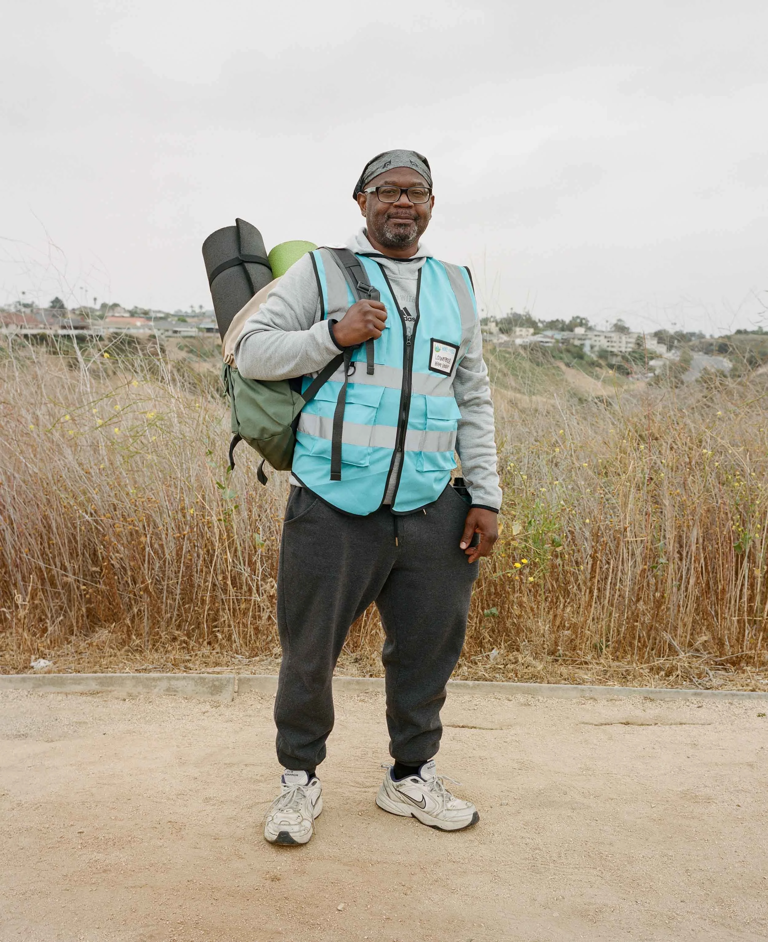 An environmental portrait of a man carrying a backpack with a sleeping mat, standing outdoors on a dirt path with dry grass and houses in the background.