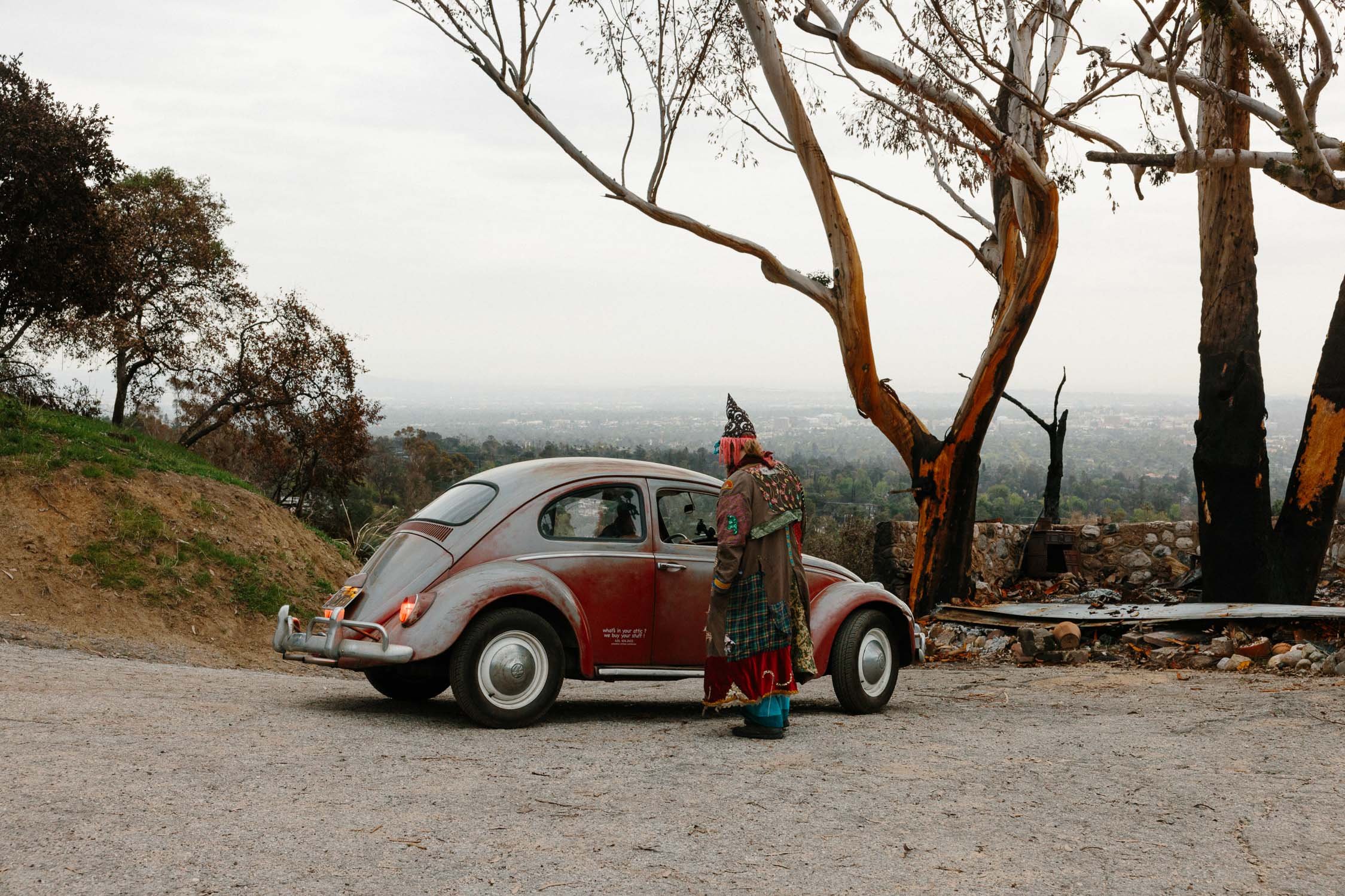 A photograph of a moment caught of a man standing near a vintage red Volkswagen Beetle on a dirt road, with burnt trees and a cityscape in the background.
