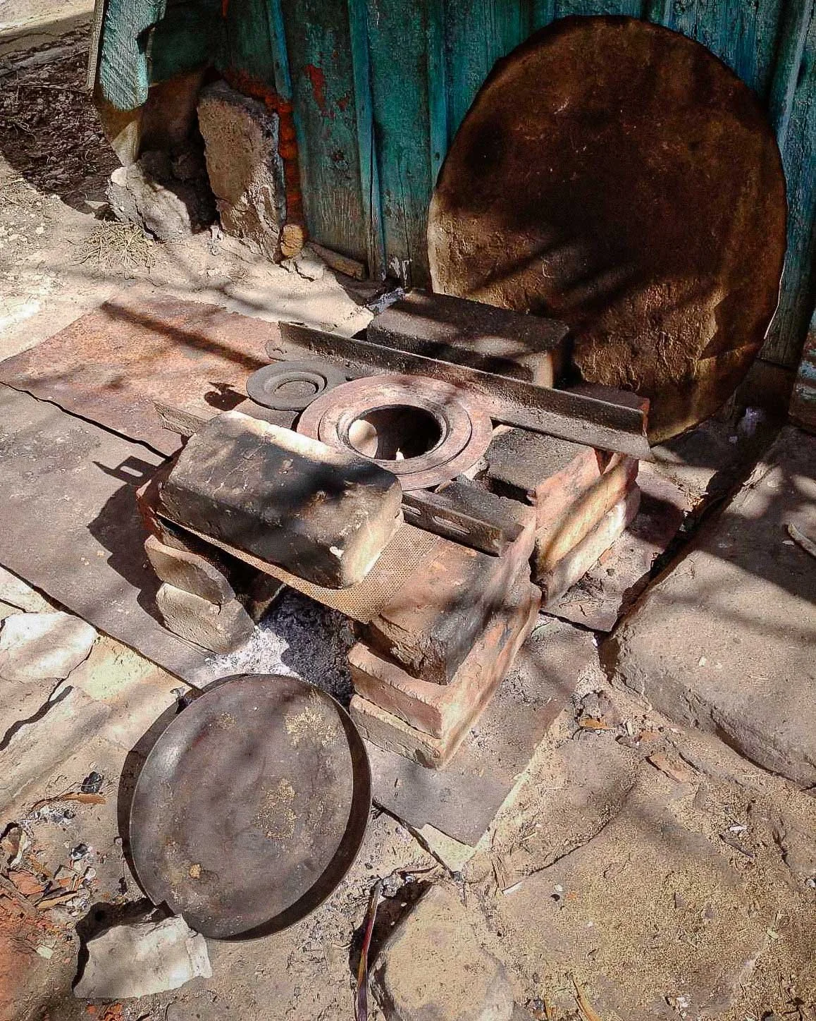 Old blacksmith forge setup with an anvil, a round circular saw blade, and a partially rusted large disk in an outdoor workshop.