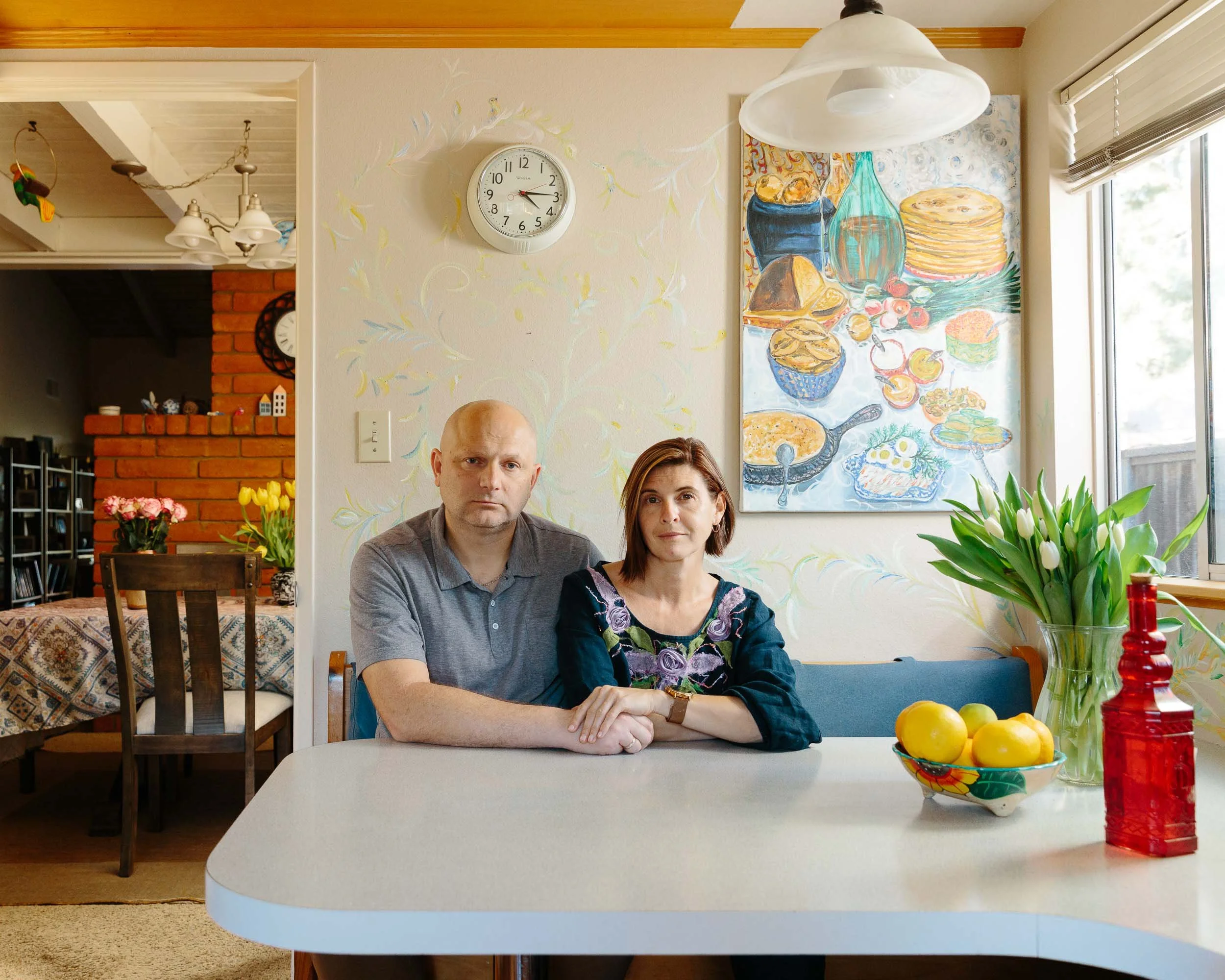 An environmental potrait of a man and woman sitting together at a dining table in a cozy kitchen with orange and white walls, a large window, potted tulips, a bowl of lemons, and colorful artwork.