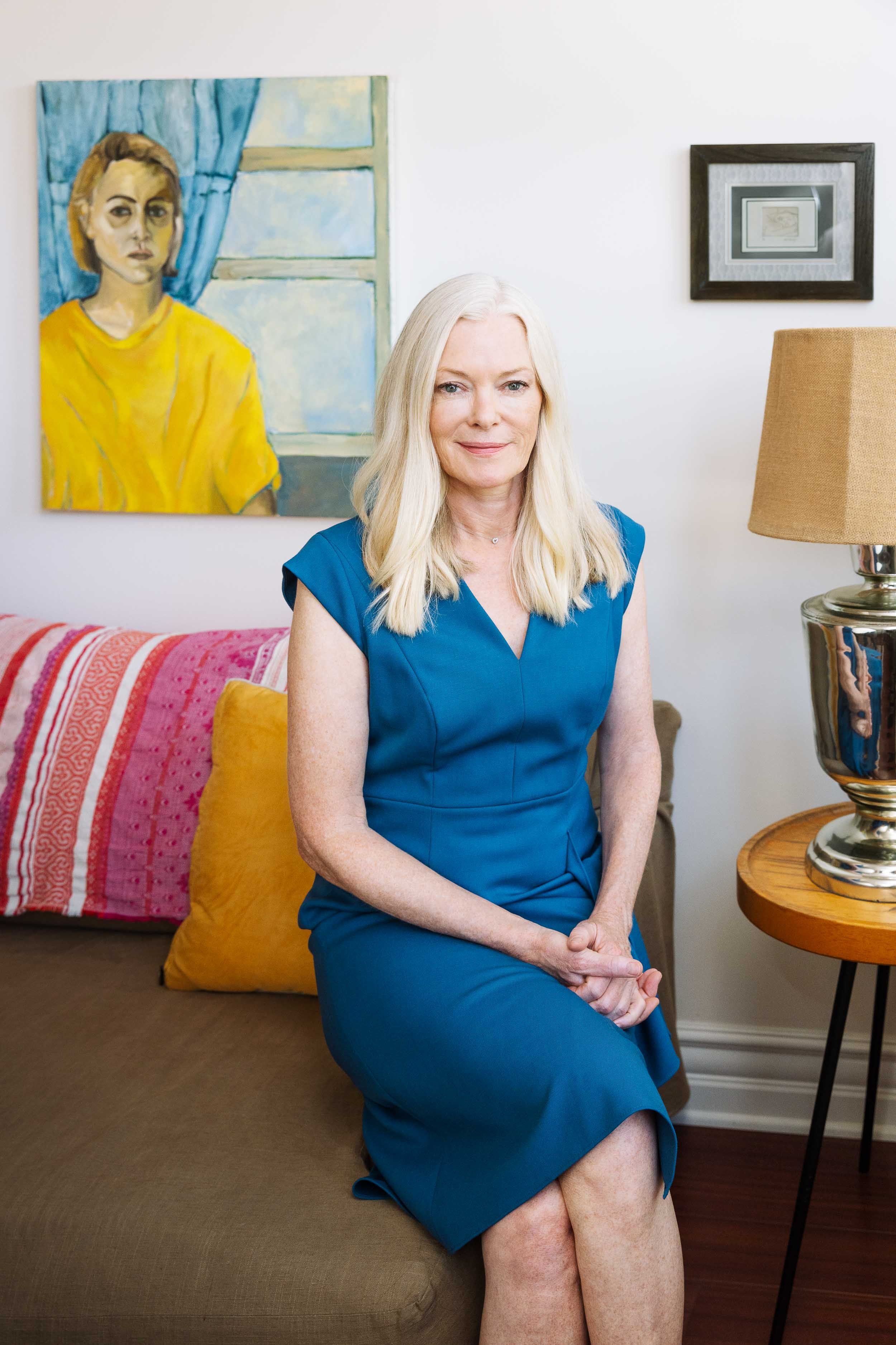 An environmental portrait of a woman with long blonde hair wearing a blue dress sits on a couch in a living room. Behind her is a colorful portrait of a woman in a yellow shirt with a window in the background.