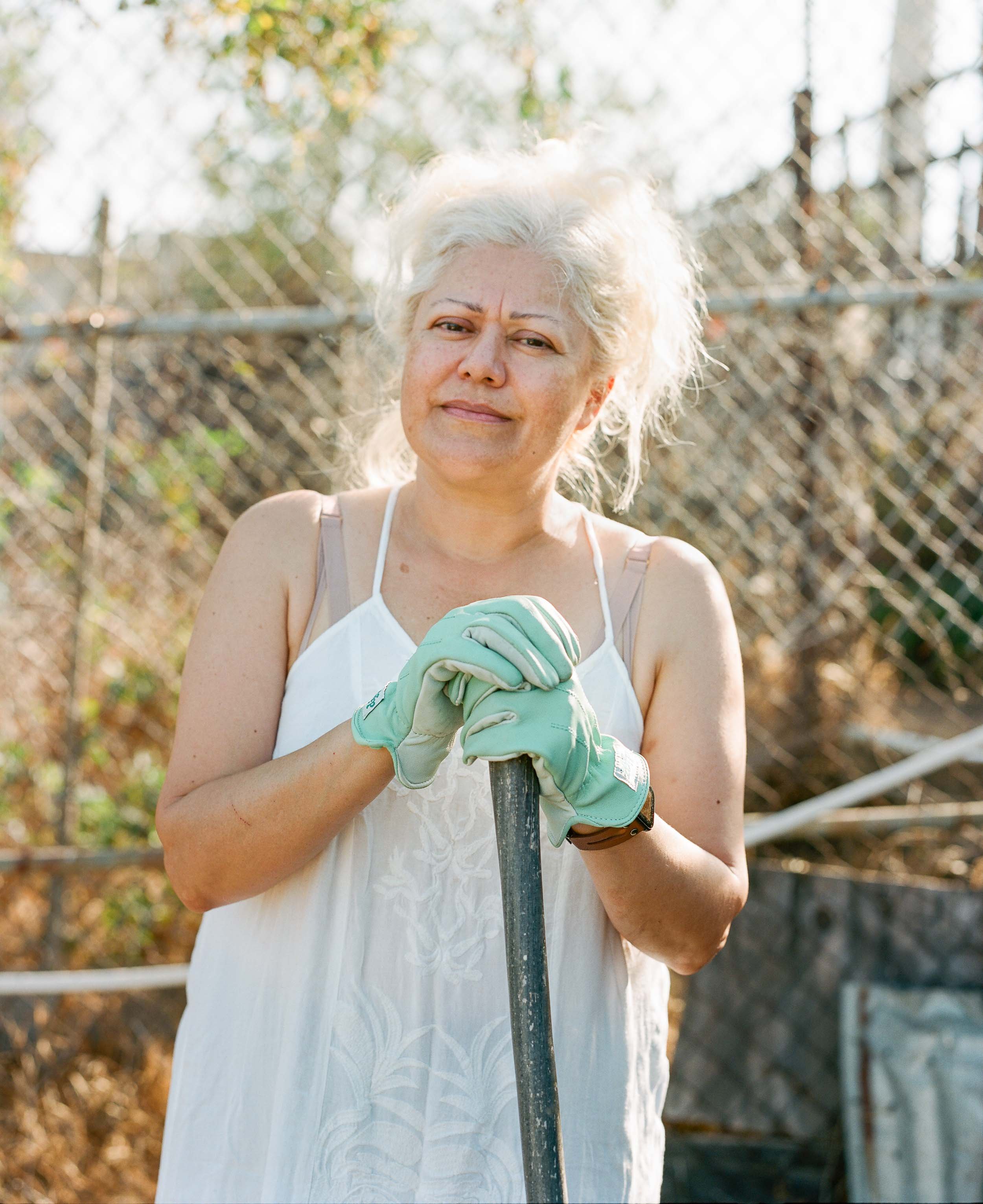 An environmental portrait of an older woman with gray hair holding a metal rake, wearing gardening gloves and a white sleeveless top, standing outdoors near a chain-link fence with trees and sunlight.