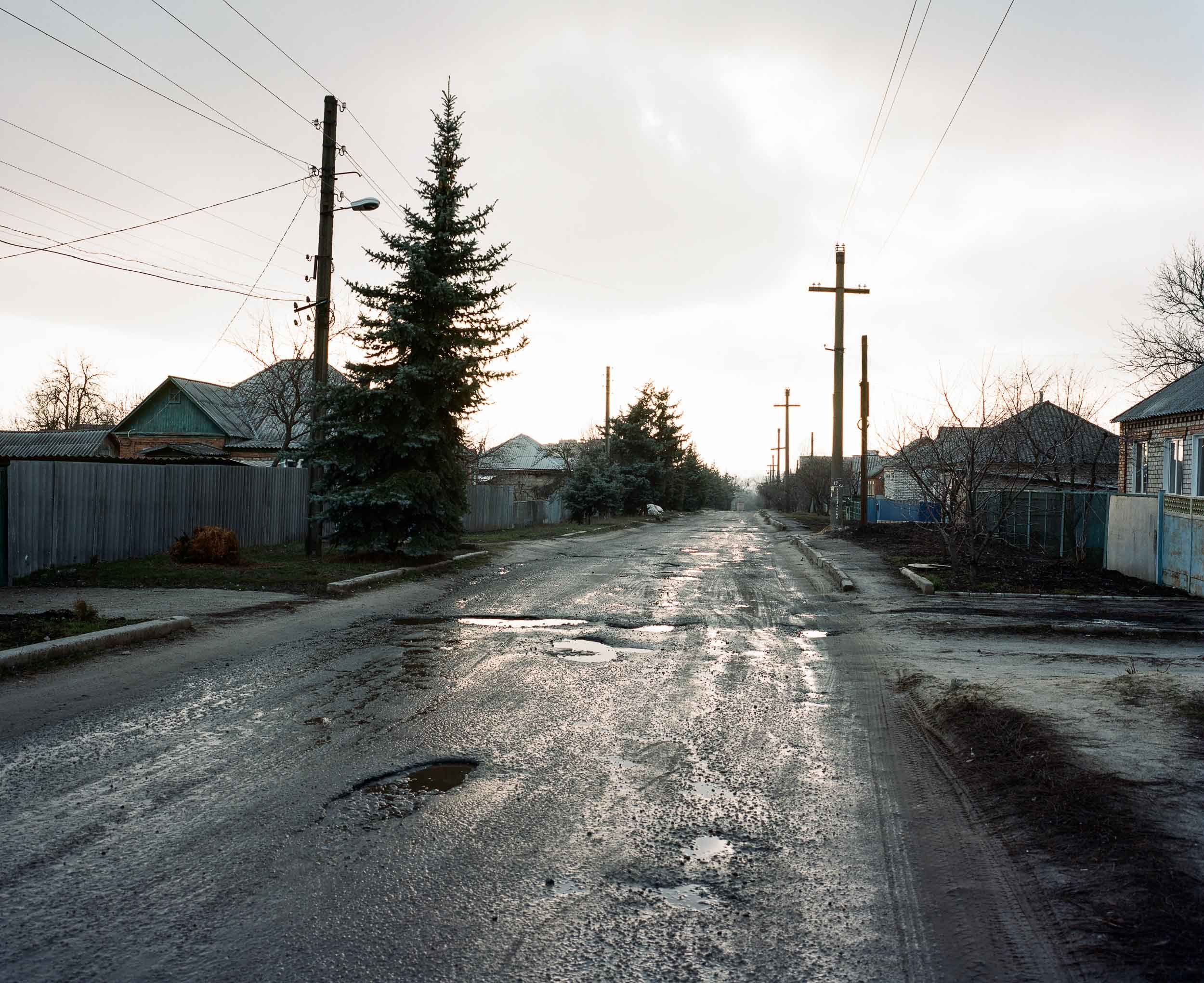 A documentary photograph of a rural street scene with muddy, wet road and puddles, lined with wooden and metal fences, residential houses, and tall pine trees, under a cloudy sky in late afternoon.