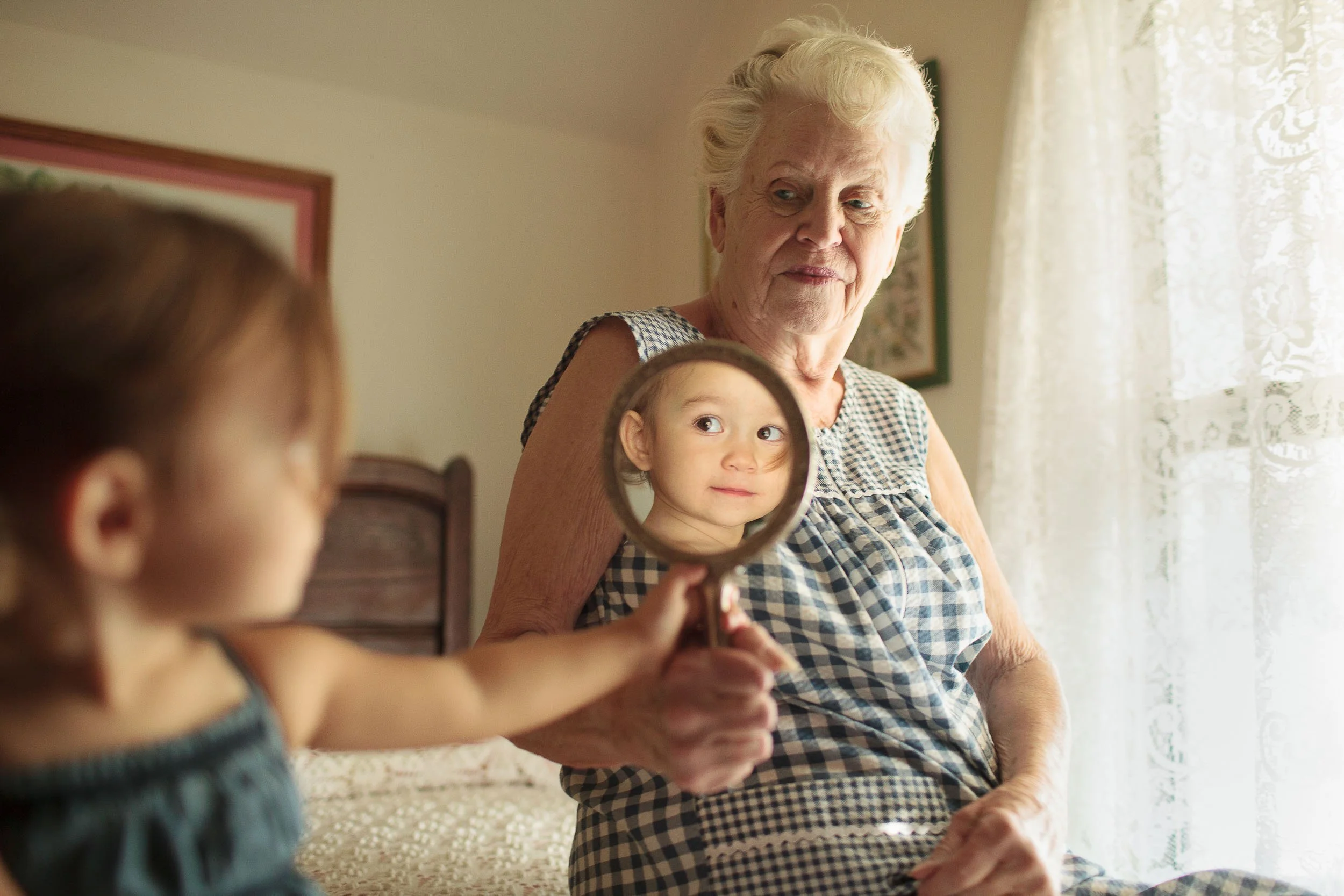 A lifestyle photograph of a young girl holding a mirror showing her reflection in front of an elderly woman in a bedroom with lace curtains.
