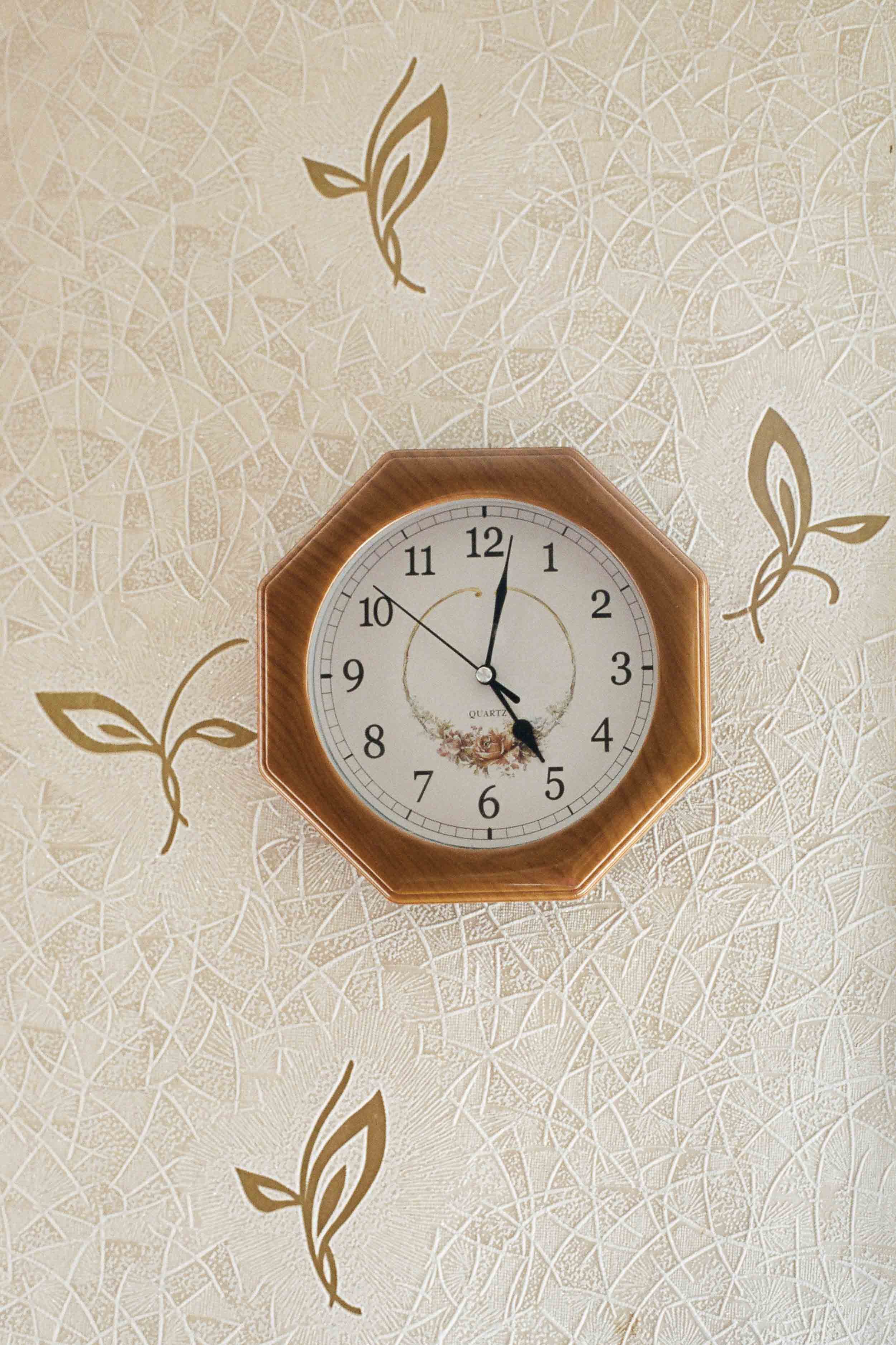 A documentary detail photograph of a round wall clock with a wooden frame on a beige wall with a textured pattern and leaf motifs, showing the time as 4:00.