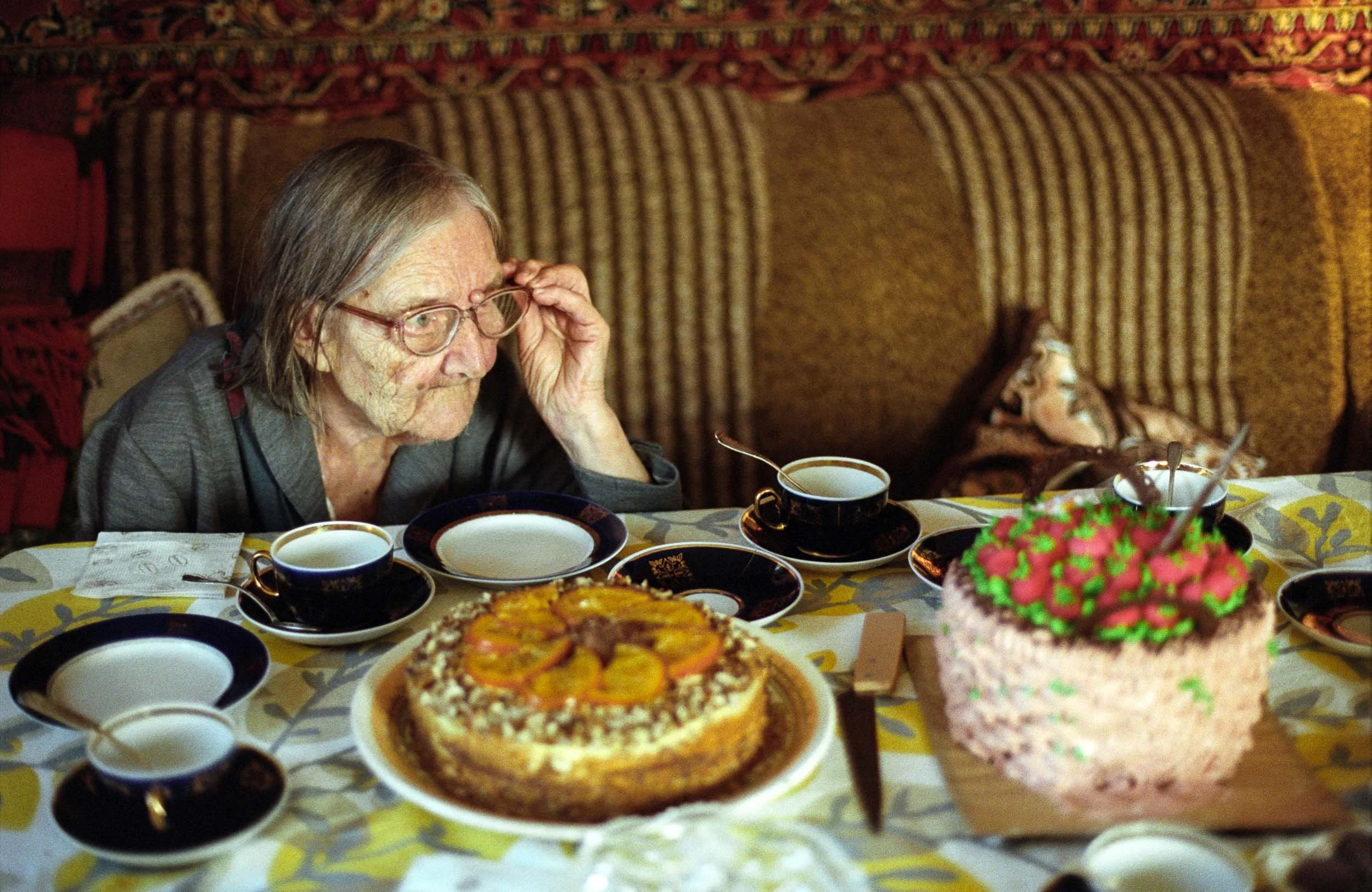 A documentary photograph of an elderly woman with glasses sitting at a table with a cake and tea cups, in a cozy room with patterned furniture.