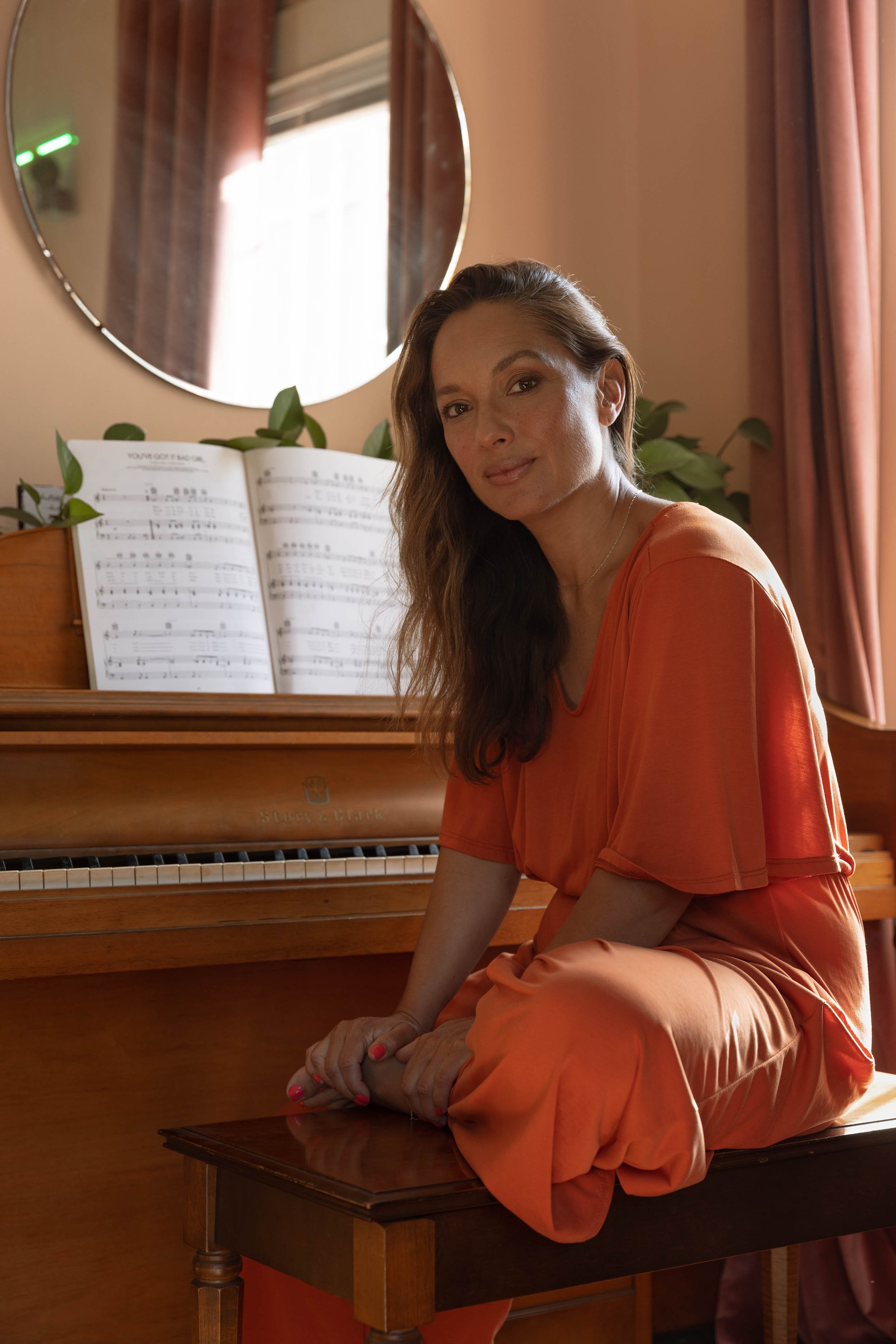 An environmental editorial portrait of a woman in an orange dress sitting on a wooden bench in front of a piano, with sheet music open on the piano, in a room with pink curtains and a round mirror on the wall.