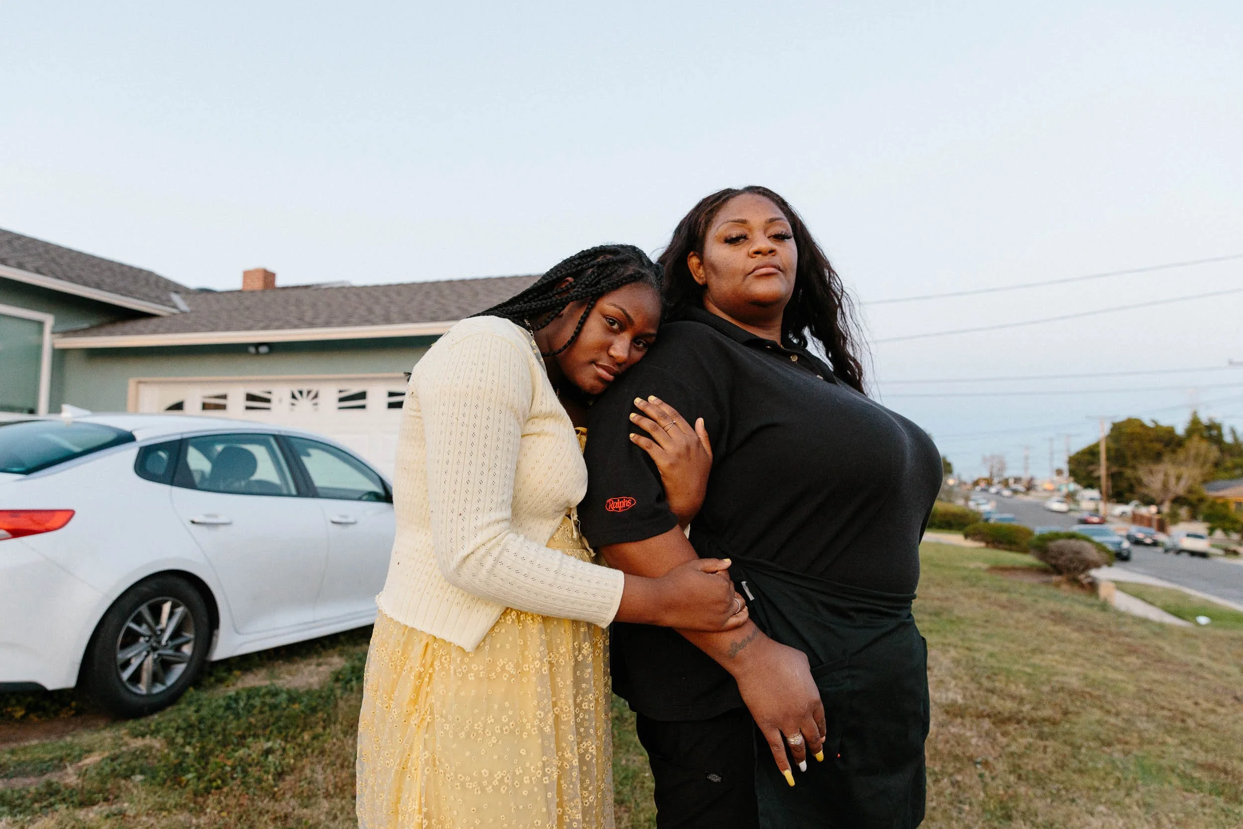 A documentary portrait of two women standing close together outdoors on a lawn, one with long dark hair wearing a black shirt and the other with braided hair wearing a yellow dress and cardigan, during daytime with a house and car in the background.