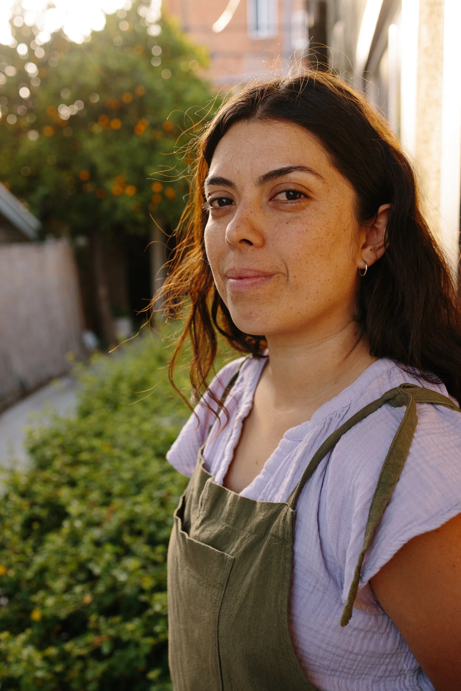 A lifestyle portrait of a woman with dark hair and freckles smiling outdoors, wearing a light purple shirt and an olive green apron, with a background of green bushes and a wooden fence, during sunset.