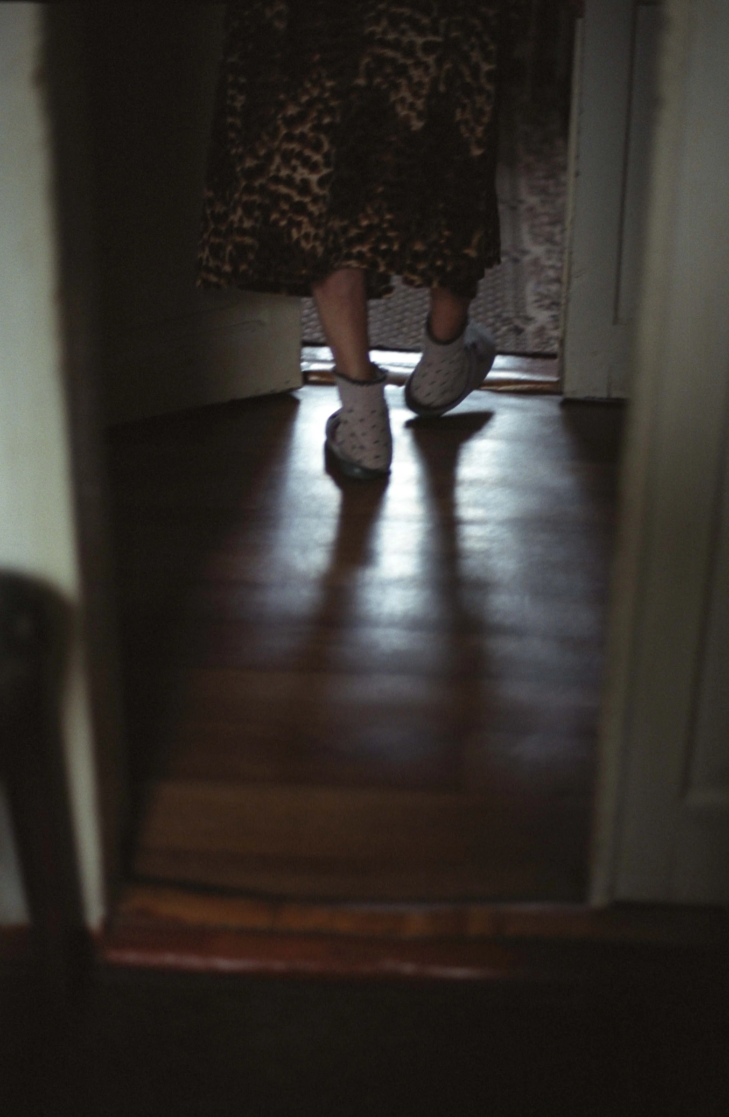 A documentary photograph of a person wearing a leopard print skirt and polka dot slippers standing in front of a mirror, with a hardwood floor visible.