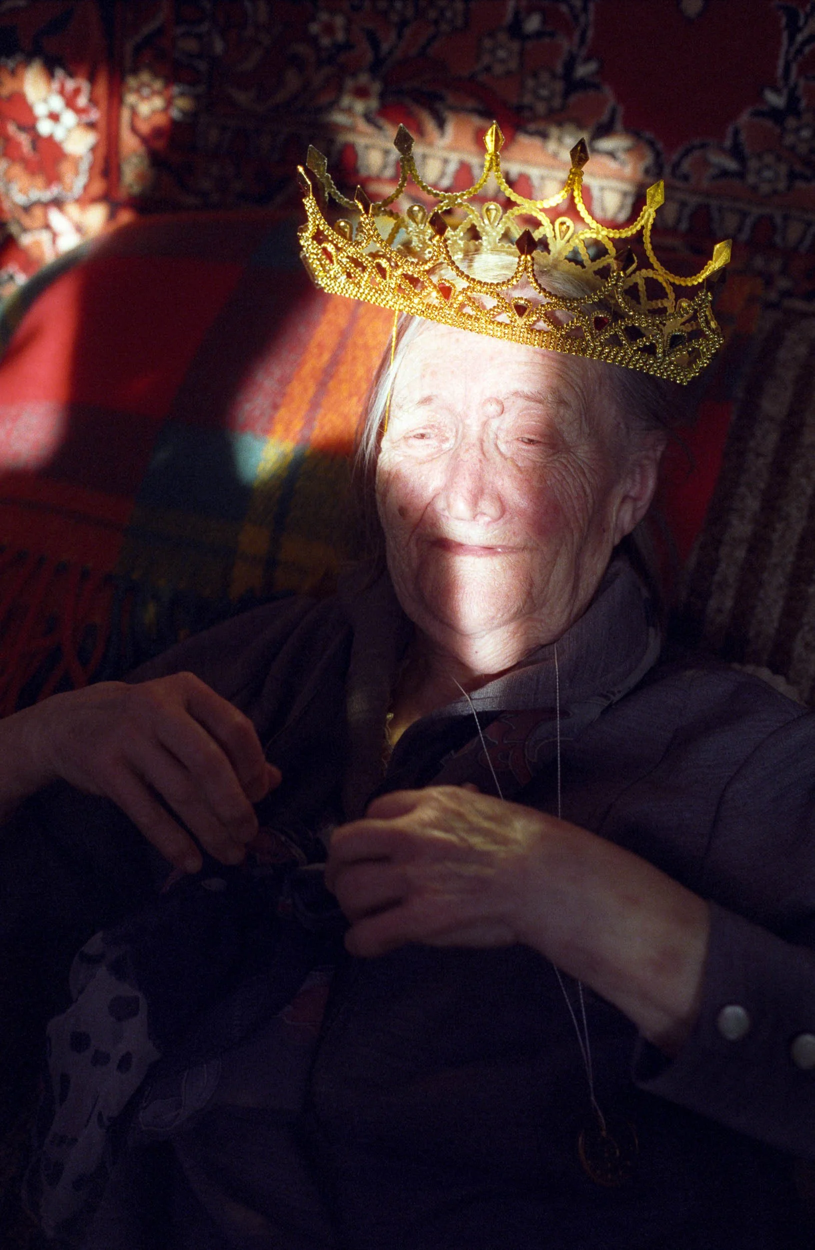 A documentary portrait of an elderly woman with a golden crown on her head, smiling gently and relaxing on a chair with a patterned cloth and shadowed sunlight on her face.