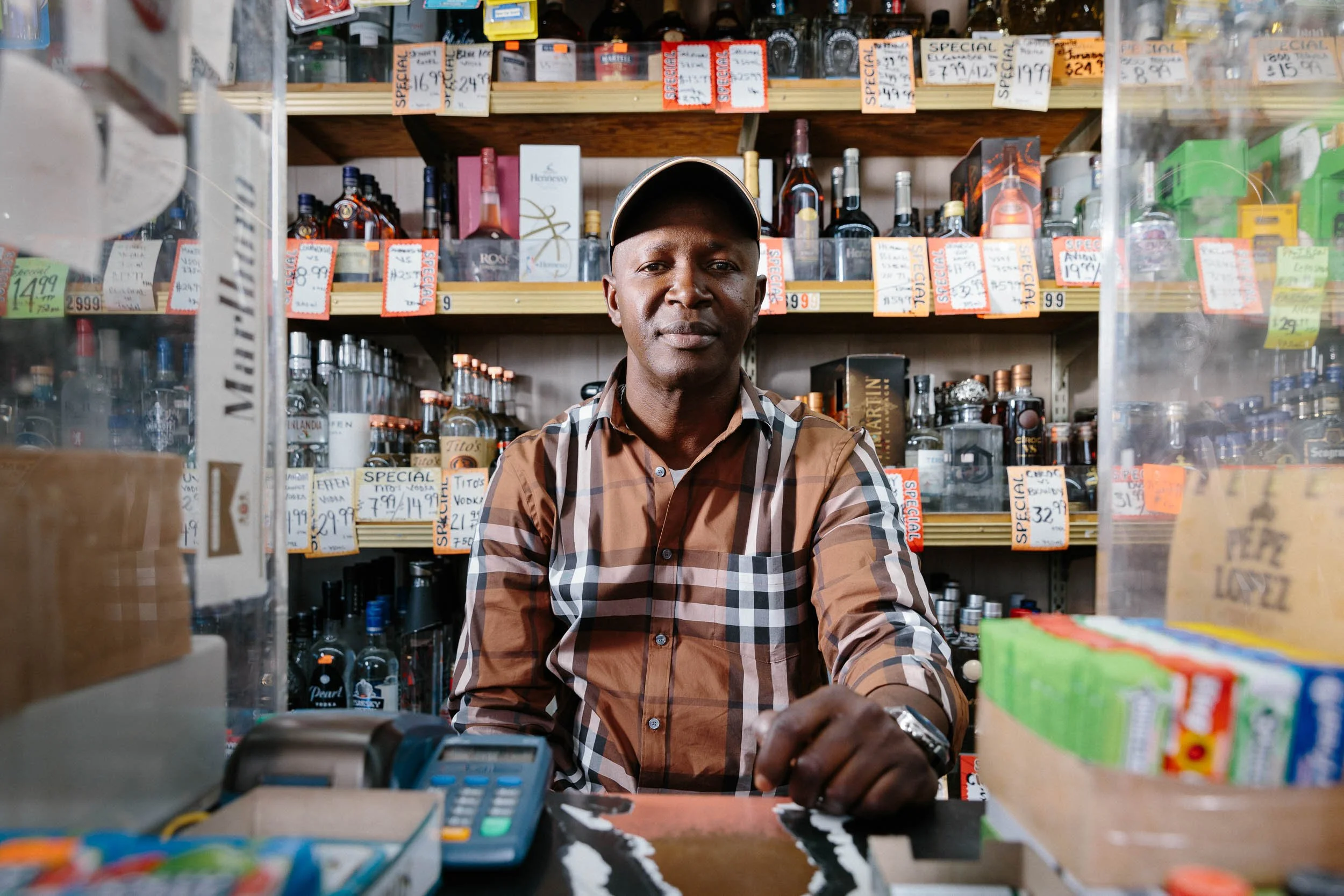 A medium environmental portrait of a man wearing a brown checkered shirt and a beige cap sitting at a liquor store counter, with shelves of alcohol bottles and sale tags in the background.