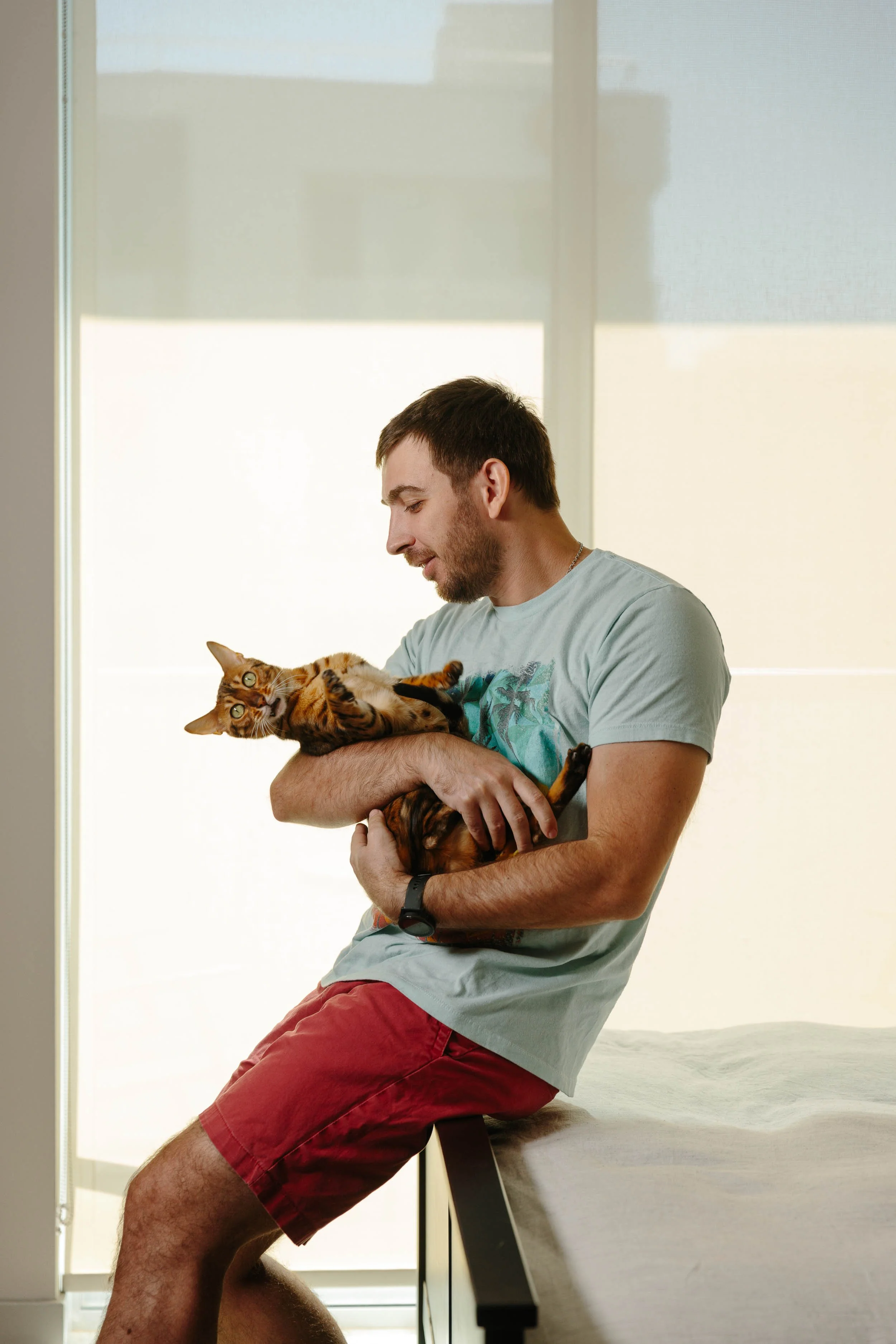 An editorial photograph of a man sitting on a bed holding a Bengal cat in his arms, with large windows in the background.