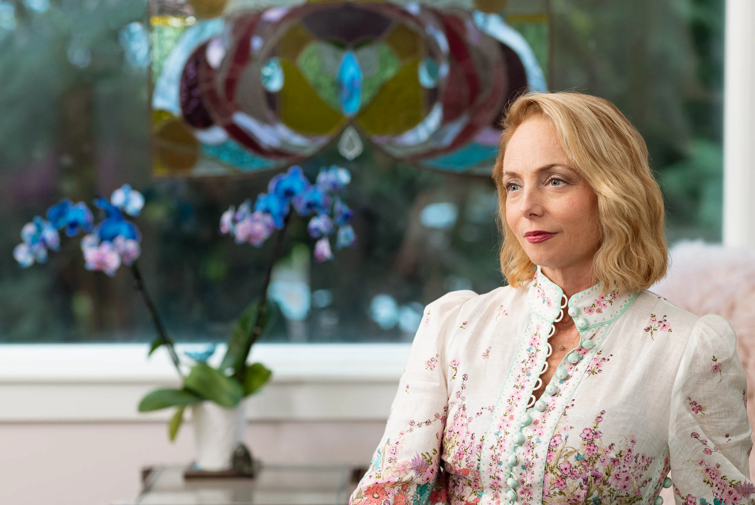 A portrait of a woman with blonde hair wearing a light-colored floral blouse sitting indoors next to a window with a blurred colorful stained glass hanging and a pot with purple orchids in the background.