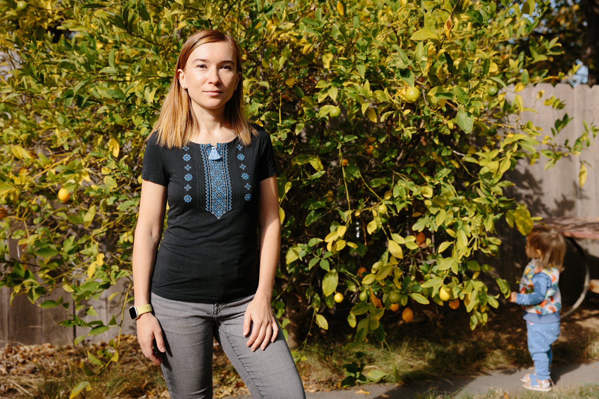 A environmental portrait of a woman with reddish-brown hair wearing a black shirt with blue embroidery and gray pants, standing outdoors in front of a lemon tree, with a young girl in a colorful jacket and blue pants picking lemons in the background.