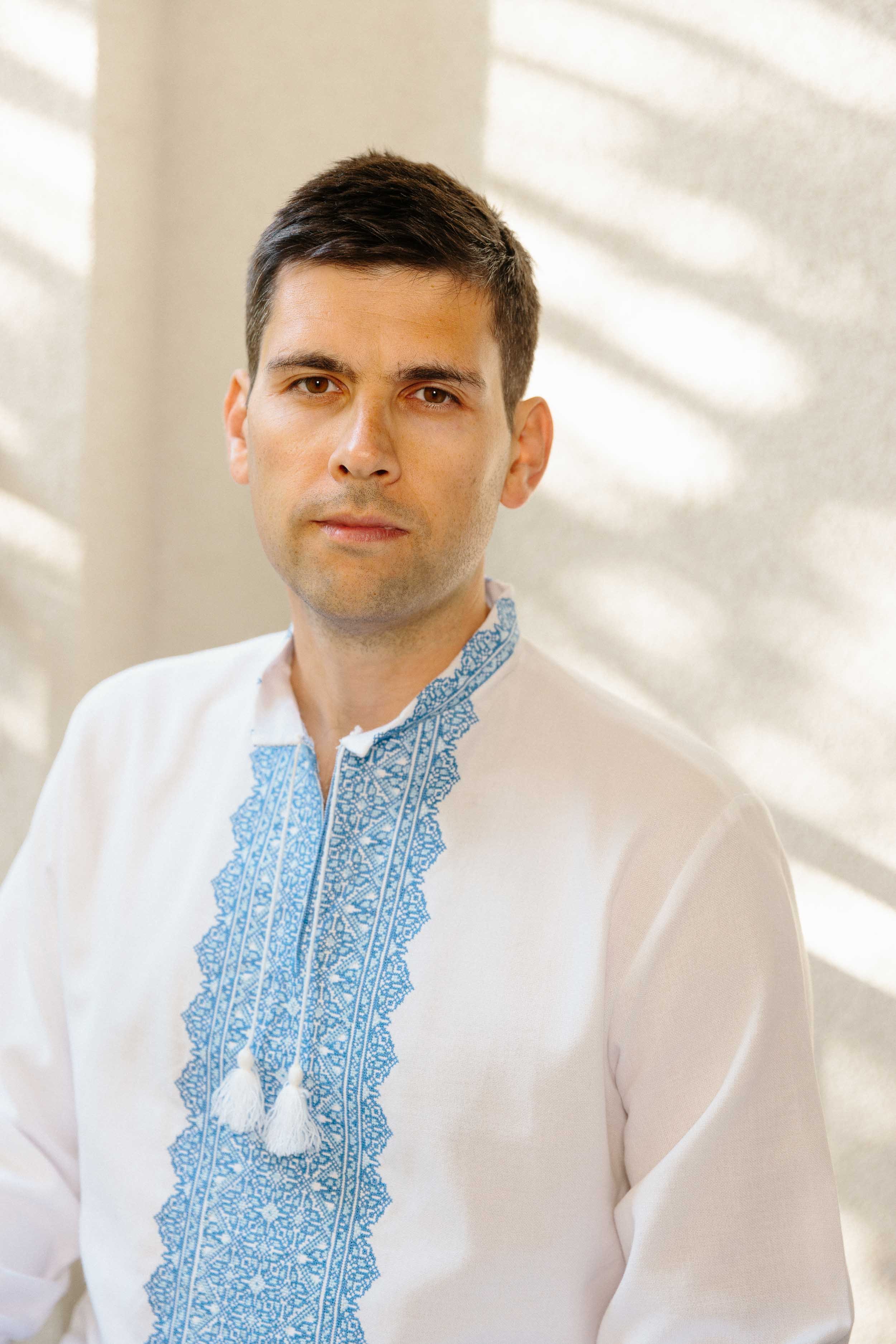 A medium environmental portrait of a young man with short dark hair and fair skin wearing traditional Ukrainian embroidered shirt with blue and white design, standing indoors with sunlight streaming through window blinds in the background.