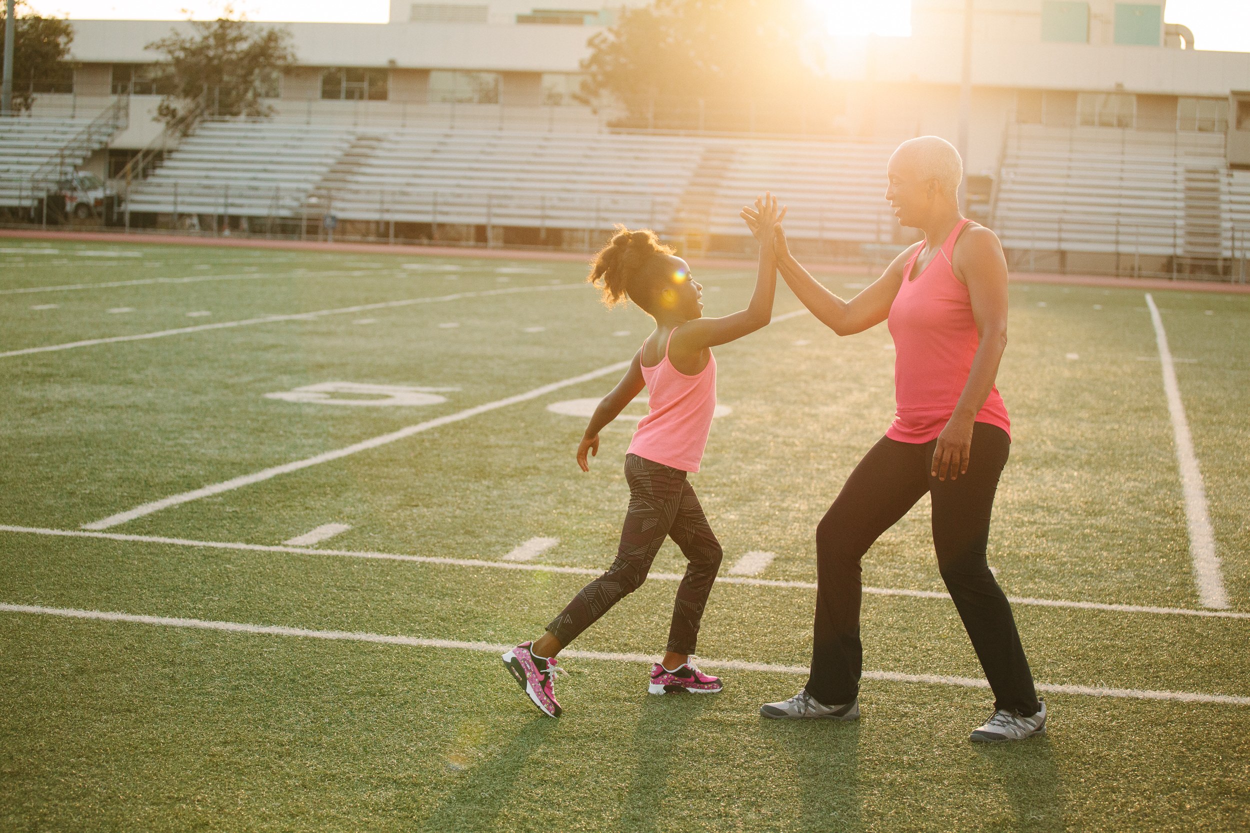 A lifestyle photograph of a young girl and an older woman wearing pink tank tops are on a football field giving each other a high-five during sunset.