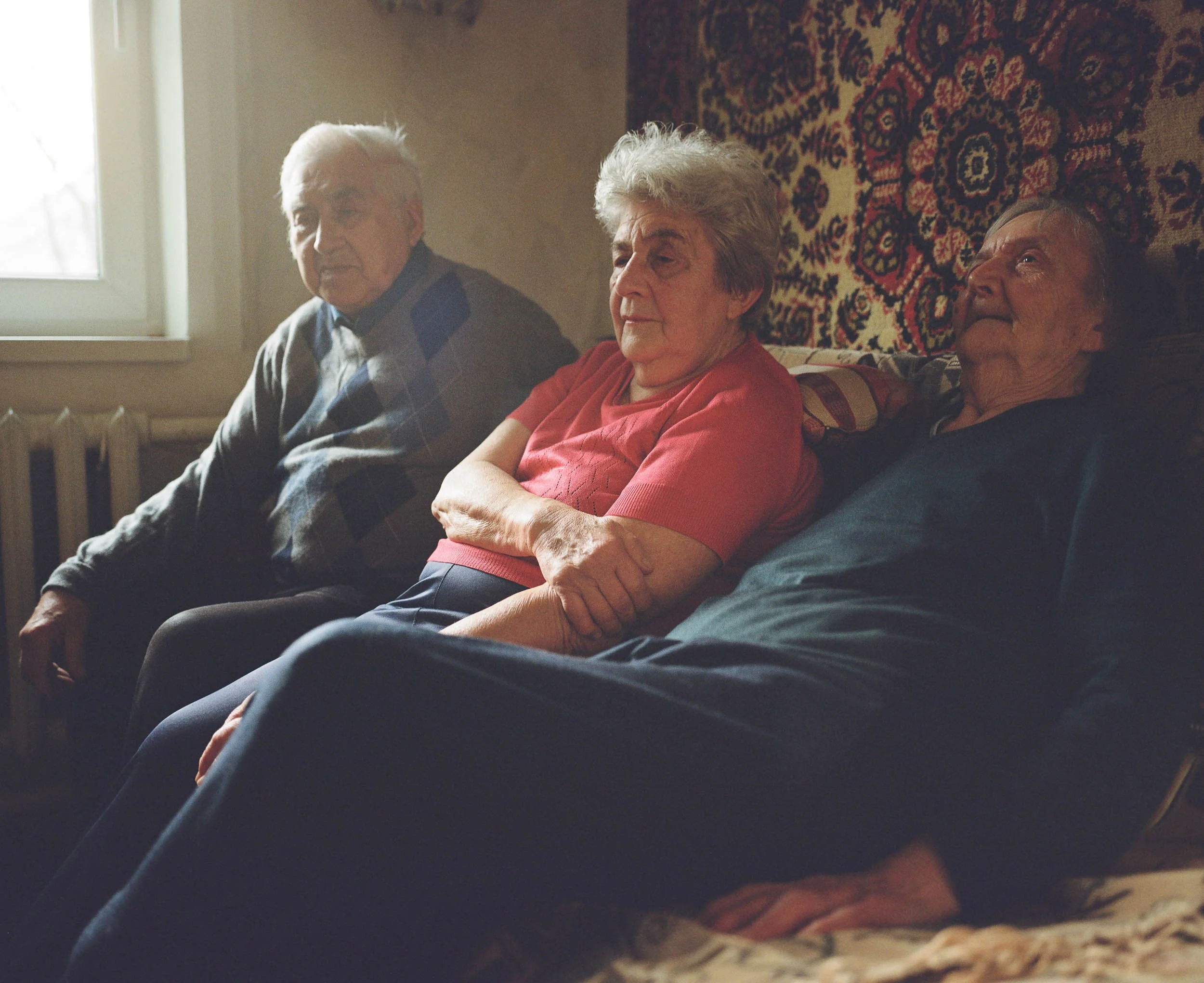 A documentary photograph of three elderly people sitting on a sofa in a living room, with one person appearing to be asleep. A window is visible in the background.
