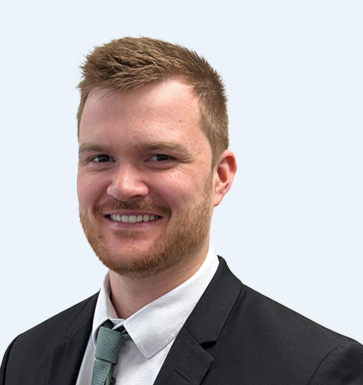 A young man with light skin, short reddish-brown hair, a beard, wearing a black suit, white shirt, and a gray tie, smiling against a light gray background.