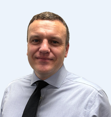 A man wearing a light blue dress shirt and a dark tie, smiling at the camera against a plain light blue background.