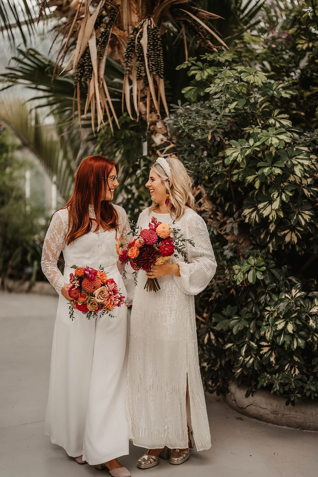 Two women in wedding dresses holding colorful bouquets, smiling and looking at each other outdoors with lush green foliage in the background.