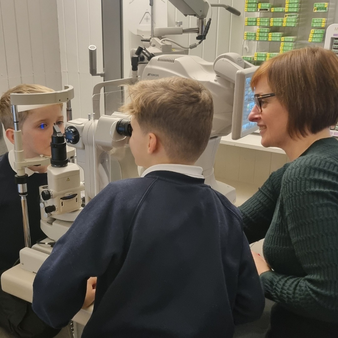 Optometrist Laura performing an eye test with a slit lamp camera on a young boy with the help of his older brother in the practice at Blink, Bridge of Allan