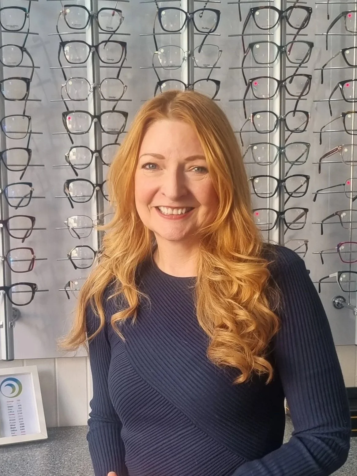 Gill Sibeth, Dispensing Optician, standing in front of a wall display of various optical frames at Blink, Bridge of Allan.