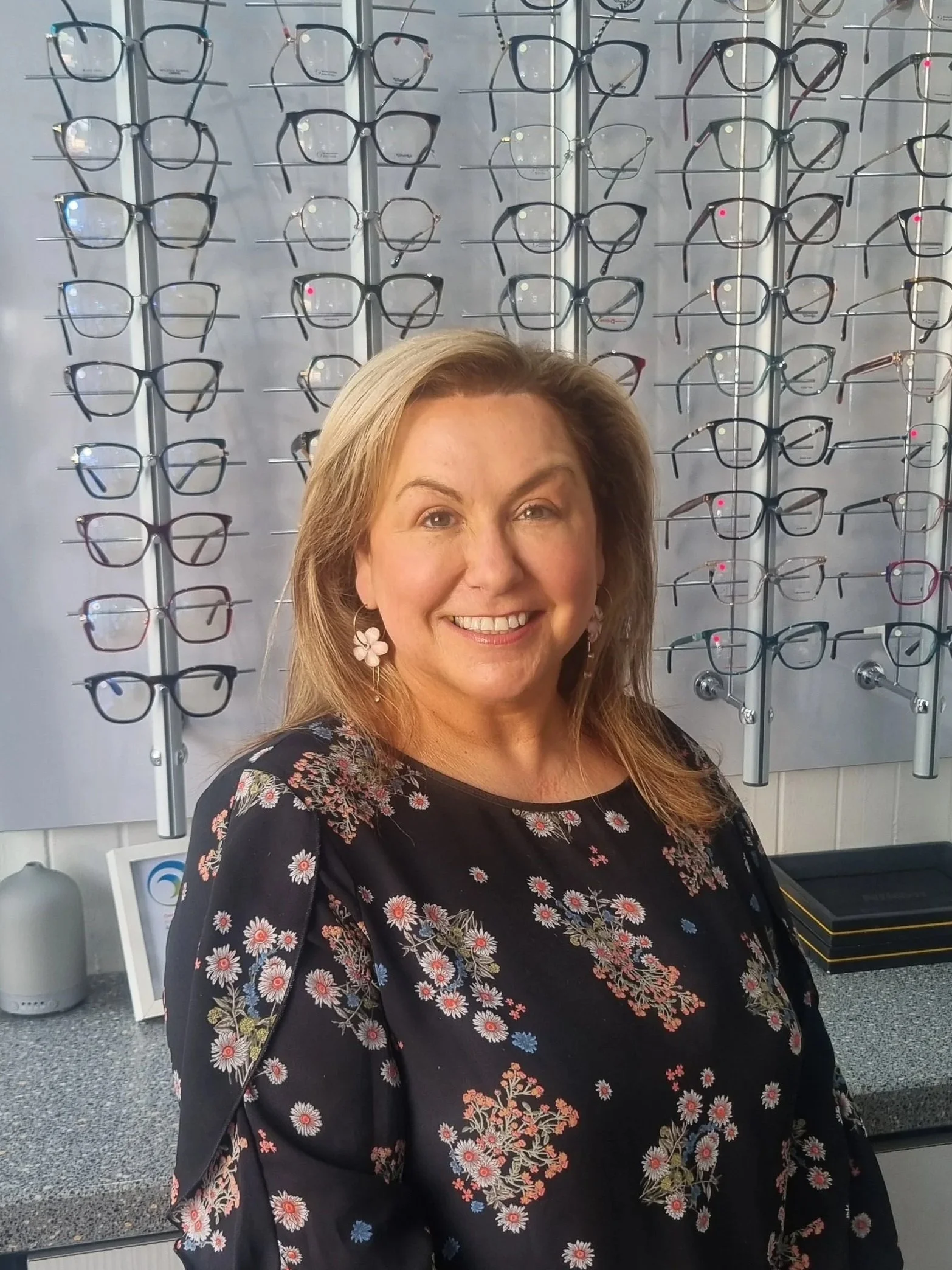 Mandy McCann Dispensing Practice Manager smiling inside Blink, Bridge of Allan, with multiple eyeglasses on display in the background.