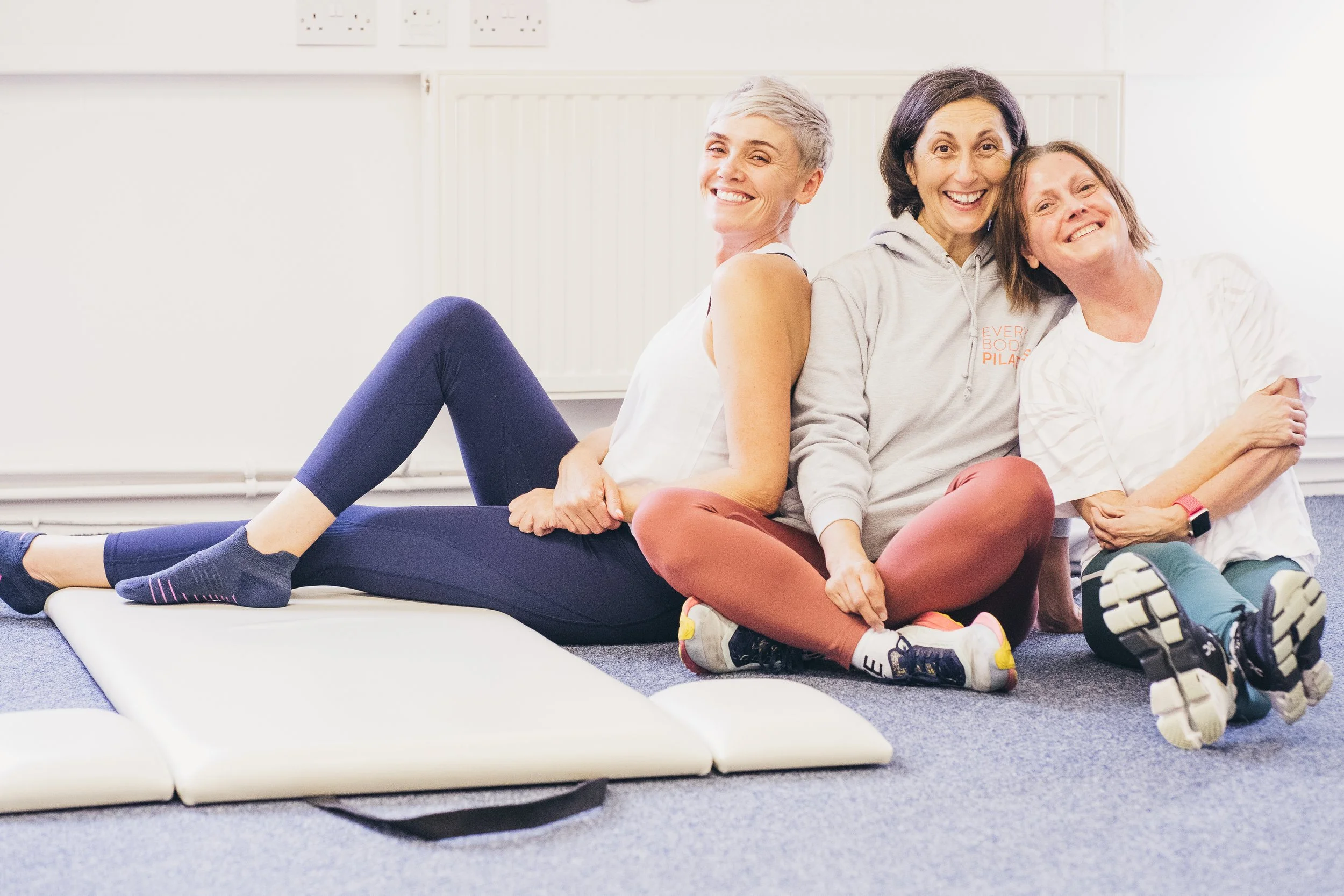 Three women sitting on the floor back-to-back, smiling, in a Pilates Studio with exercise mats on the floor.