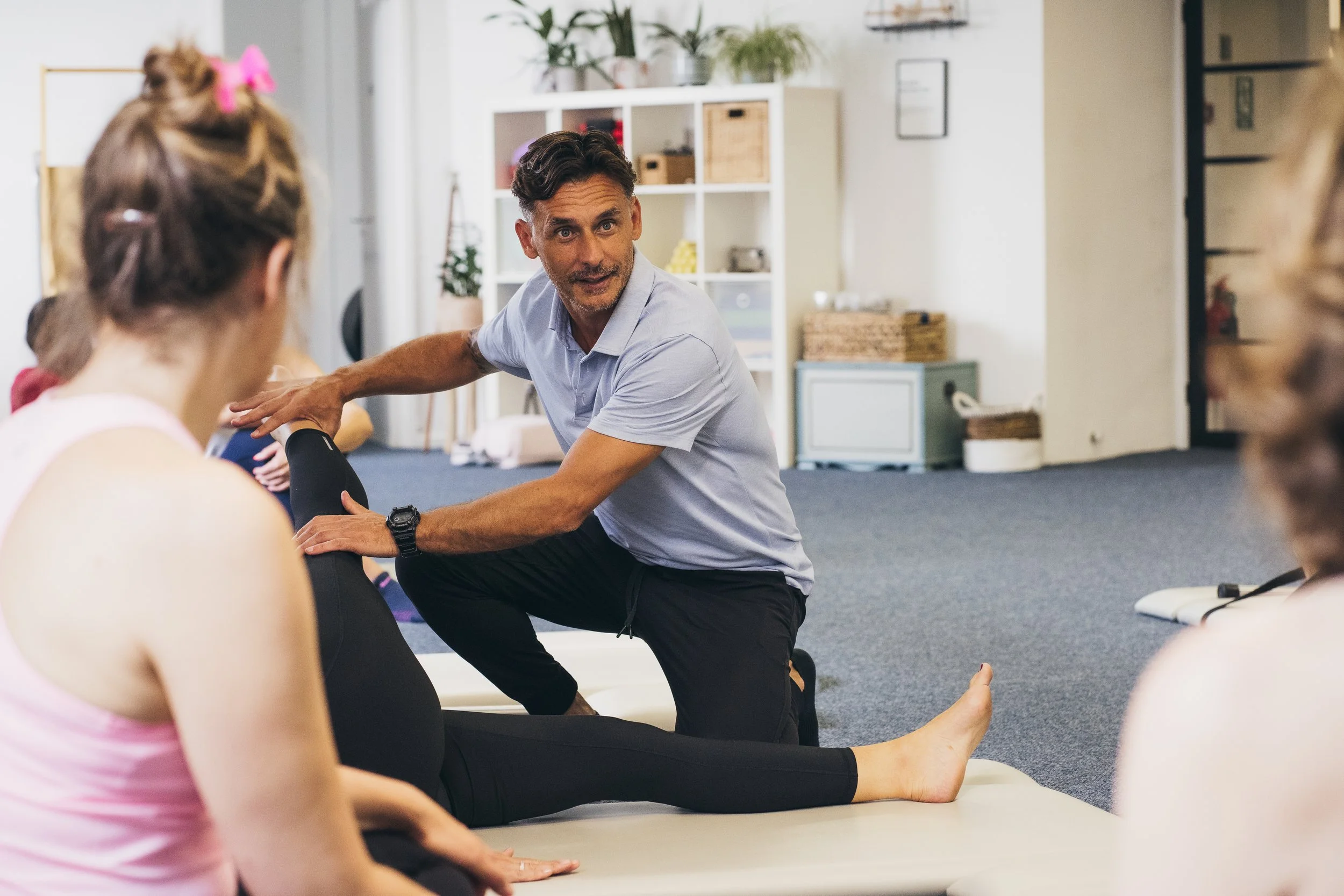 A man in a light blue shirt is demonstrating an exercise technique to a group of female Pilates Teachers seated on a mat. He is supporting a woman’s leg as she stretches. The setting is a Pilates Studio workshop inside a room with shelves and plants.