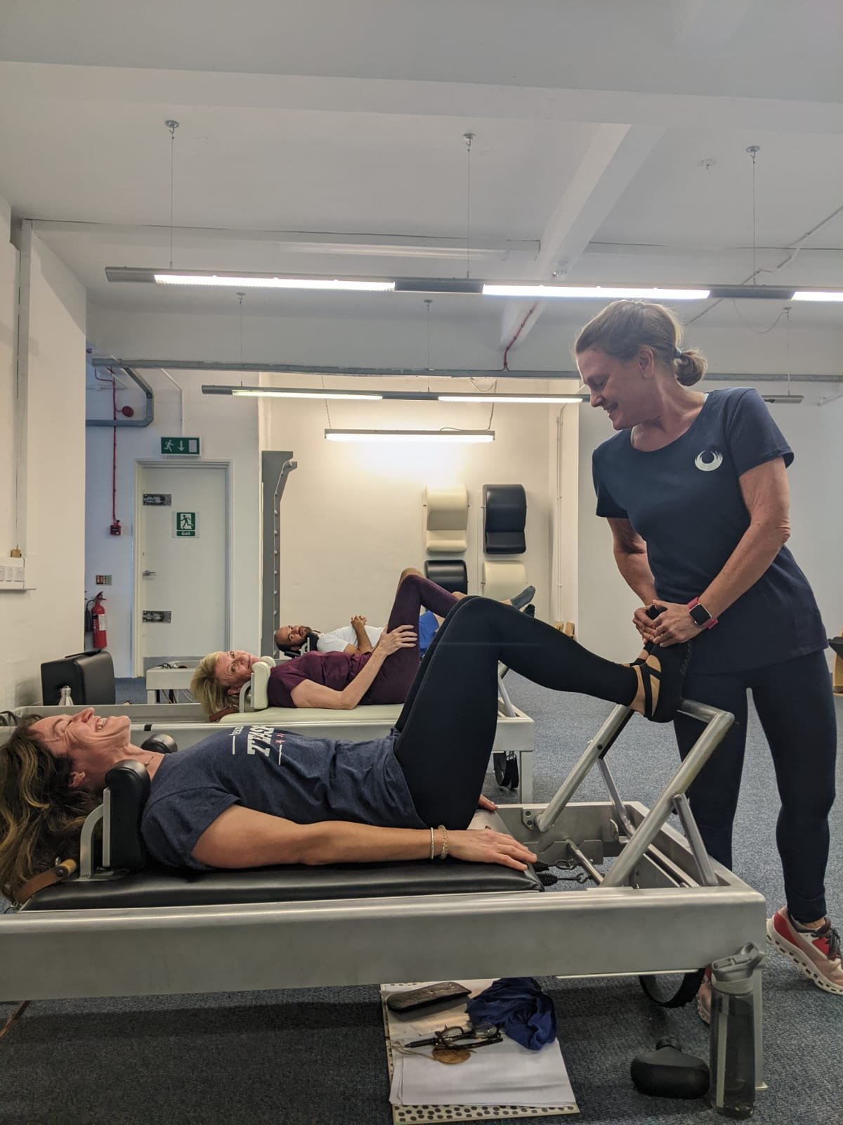 A woman lying on a Classical Pilates Reformer while exercising under guidance from a Classical Pilates Studio. Two other clients are exercising in the background.