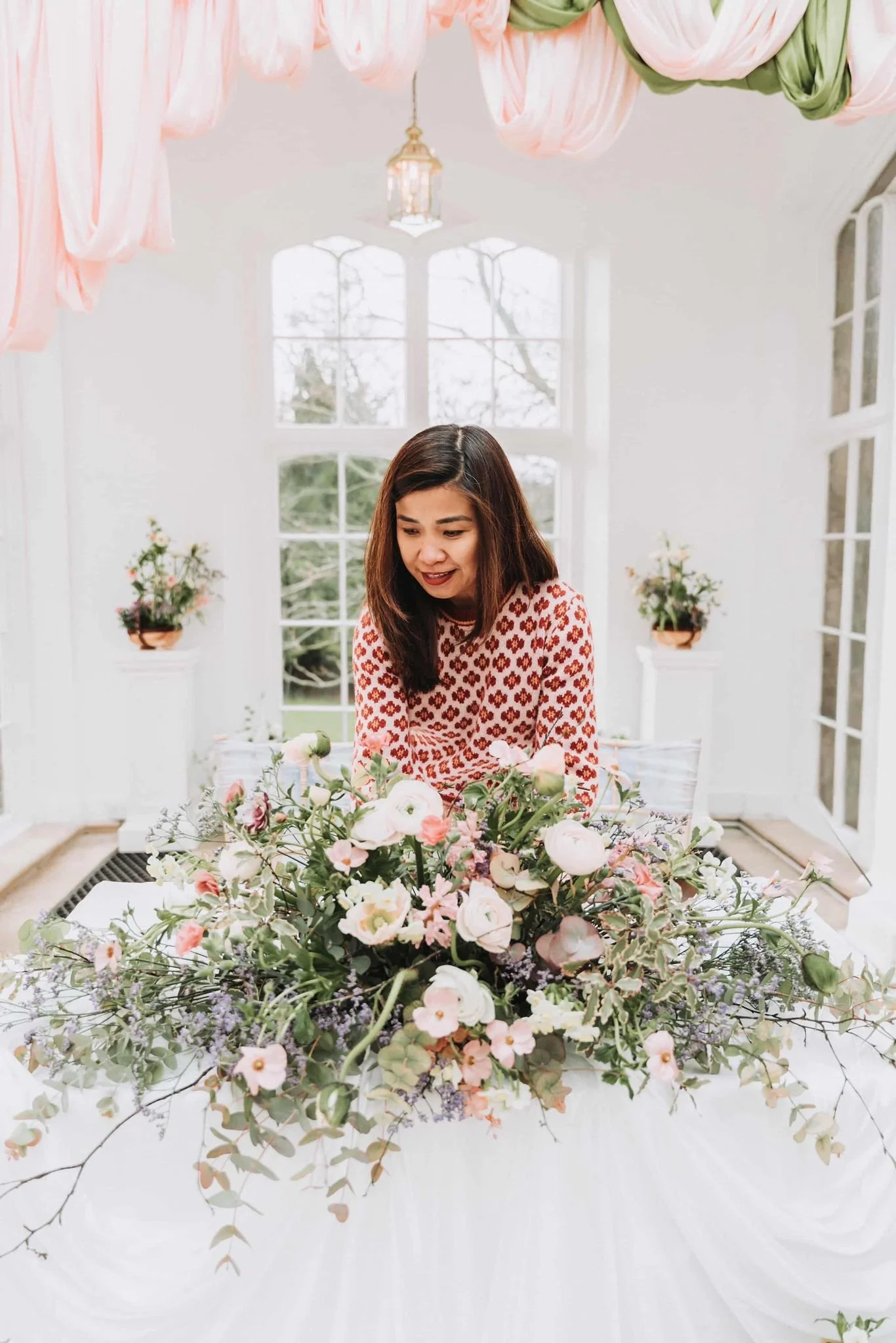 Woman arranging a large floral centerpiece with pink, white, and purple flowers in a brightly lit room with large windows and pink and green drapery hanging from the ceiling.